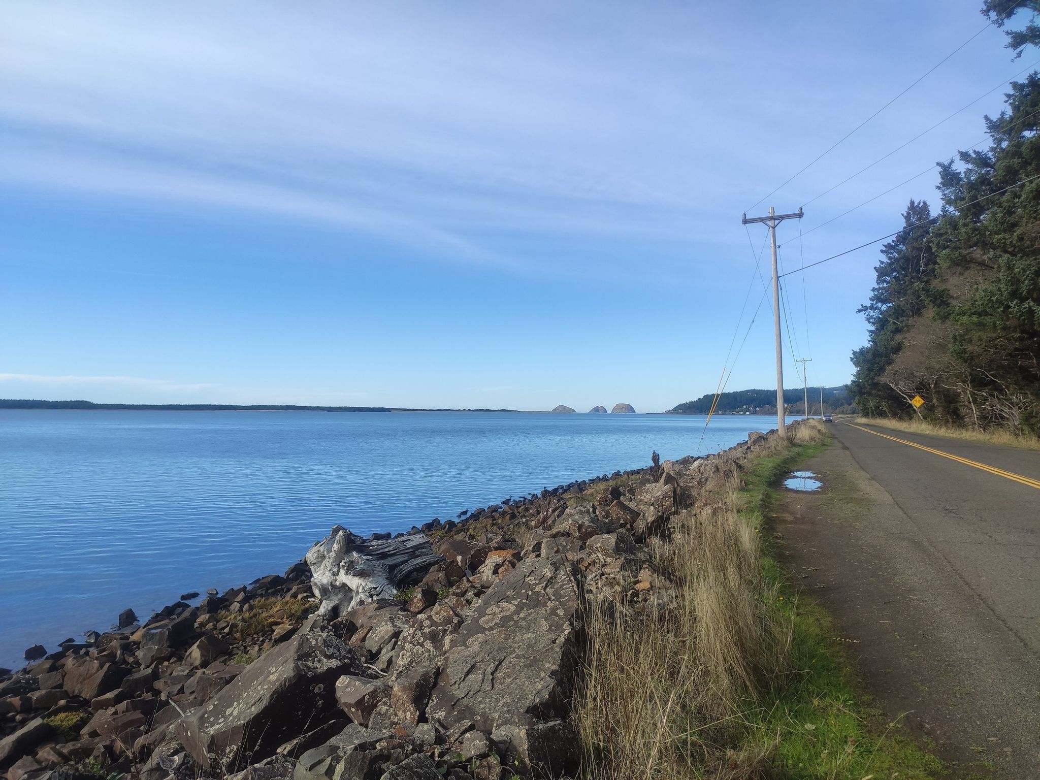 Calm blue water next to a rocky shore and a road under a clear sky. - Netarts Bay Drive – Pleasant Valley loop from Netarts