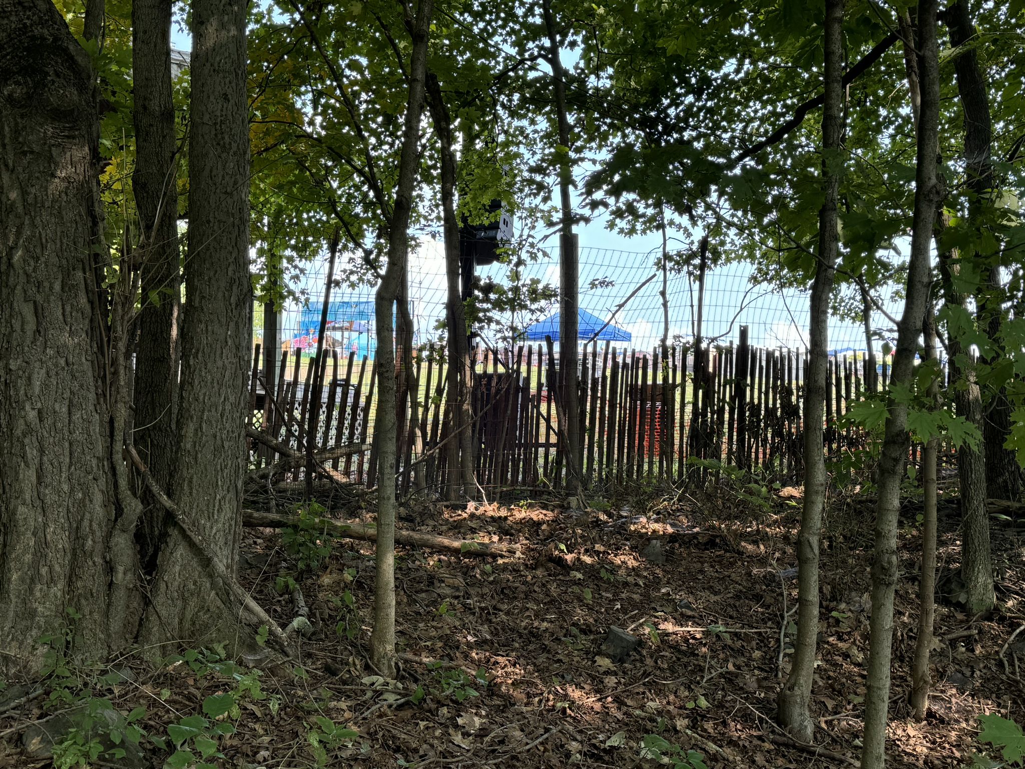 Wooden fence and trees partially obscuring distant blue structures. - Perkiomen Bicycles – Steep Climb on Perkiomen Trail loop from Lower Salford Township