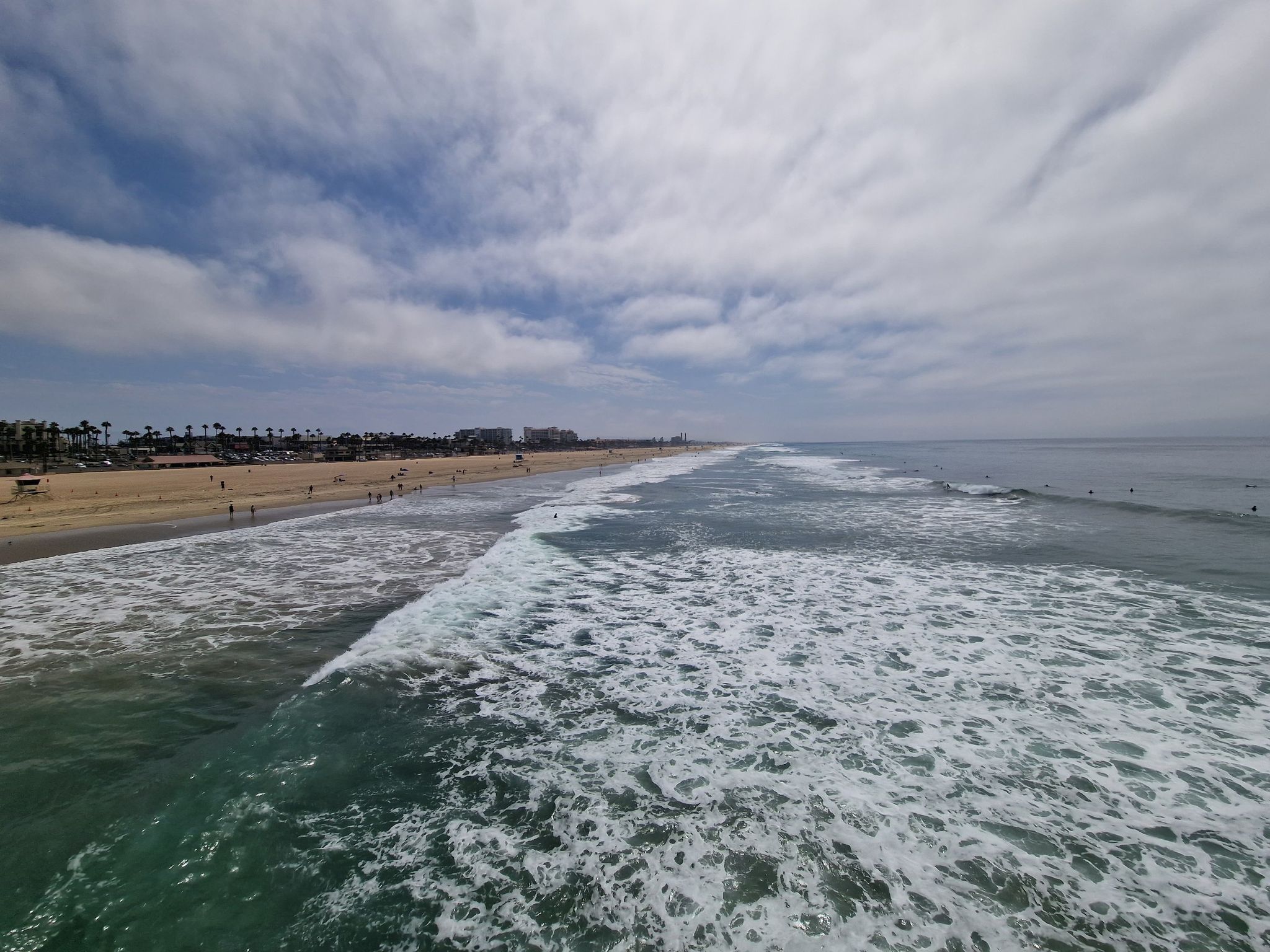 Ocean waves crashing onto a wide sandy beach with distant buildings under cloudy sky. - Huntington Beach Pier – Huntington City Beach loop from Huntington State Beach