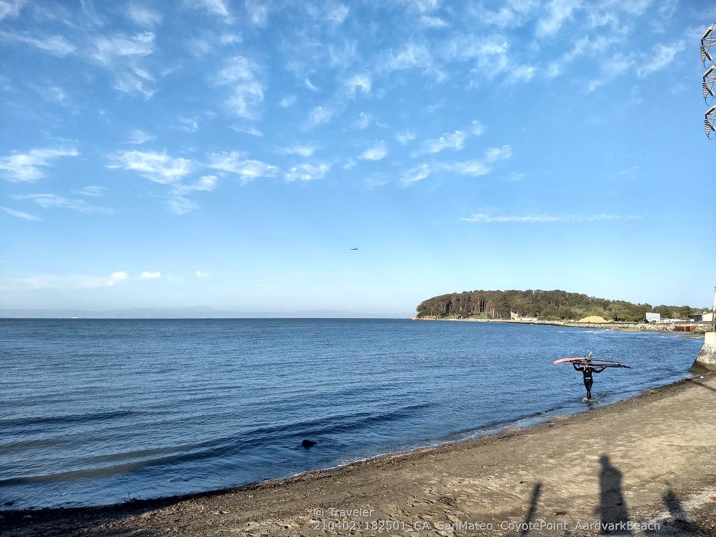 Person carrying a paddleboard walks along a sunny beach next to calm water. - Running loop from Coyote Point Recreation Area