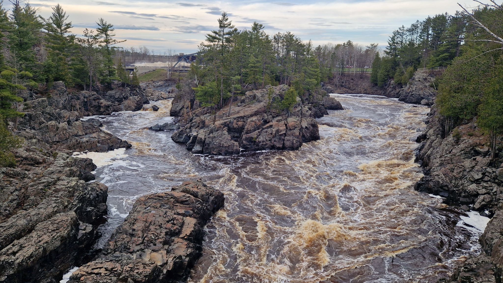 Turbulent, brown river flowing through a rocky, forested landscape under an overcast sky. - View of St. Louis River Gorge – Trail cut through the rock loop from Norton Park