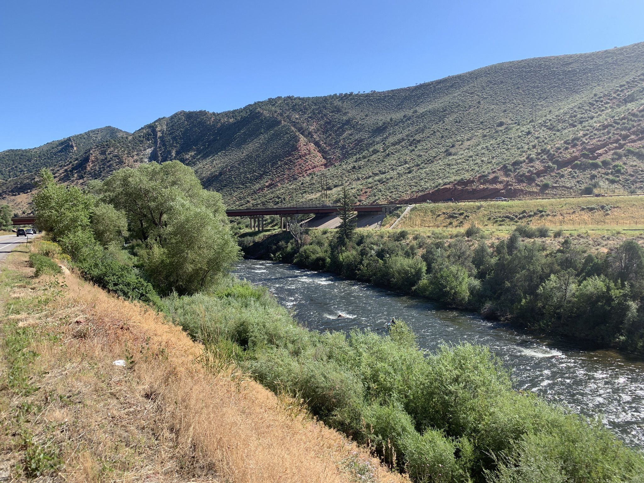 A wide river flows past green mountains and a bridge under a clear sky. - Eagle Valley Trail