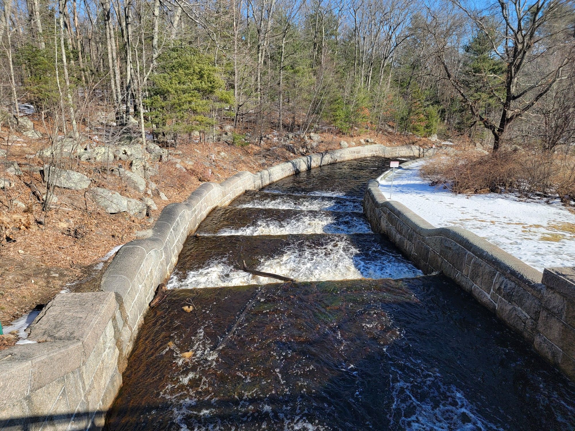 Stepped concrete channel with flowing water, surrounded by bare trees and snow. - Running loop from Hopkinton State Park