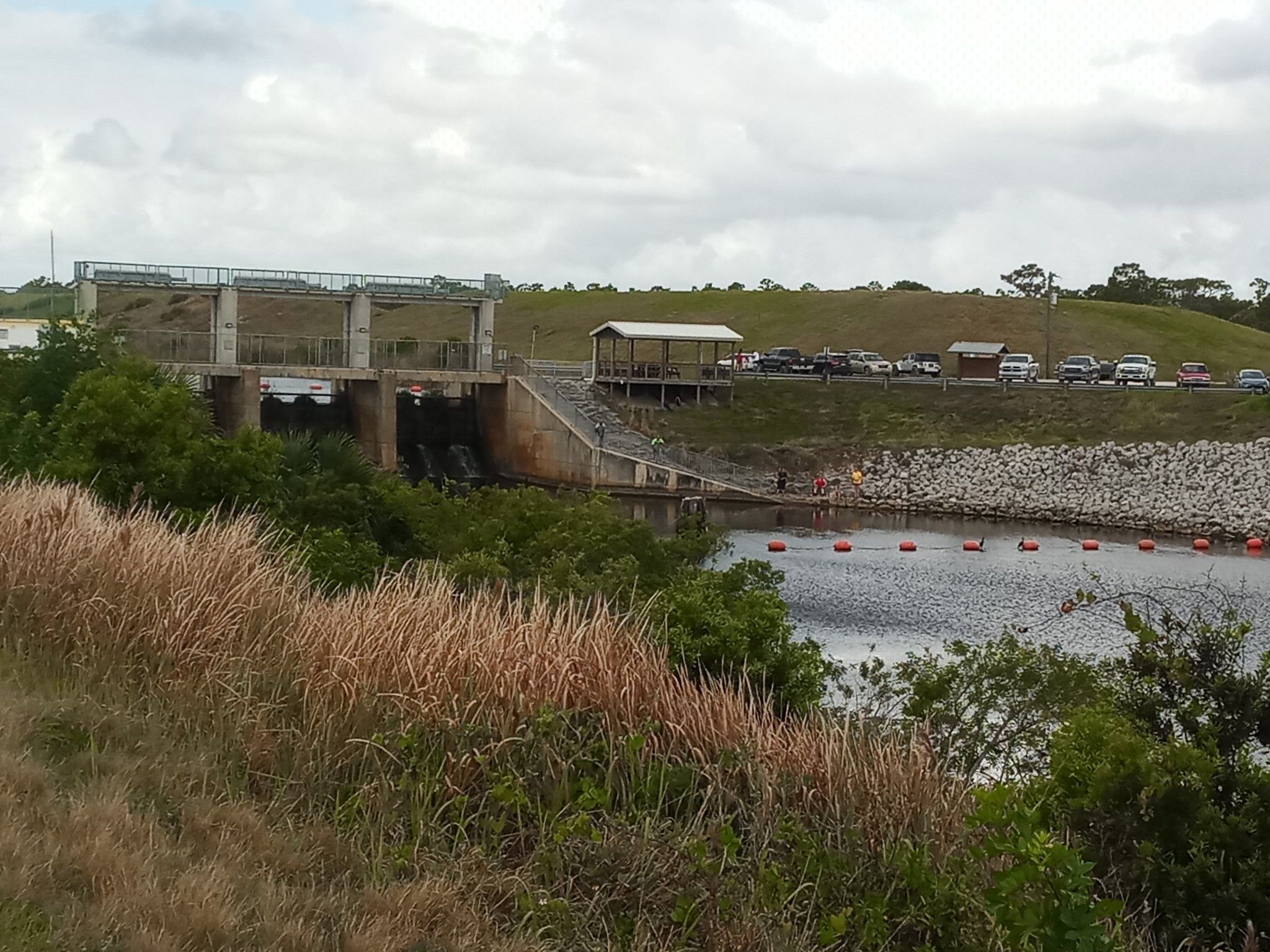 Overcast view of a dam, water with buoys, and people by a rocky bank. - St. Sebastian Southeast Quadrant Blue Trail Loop
