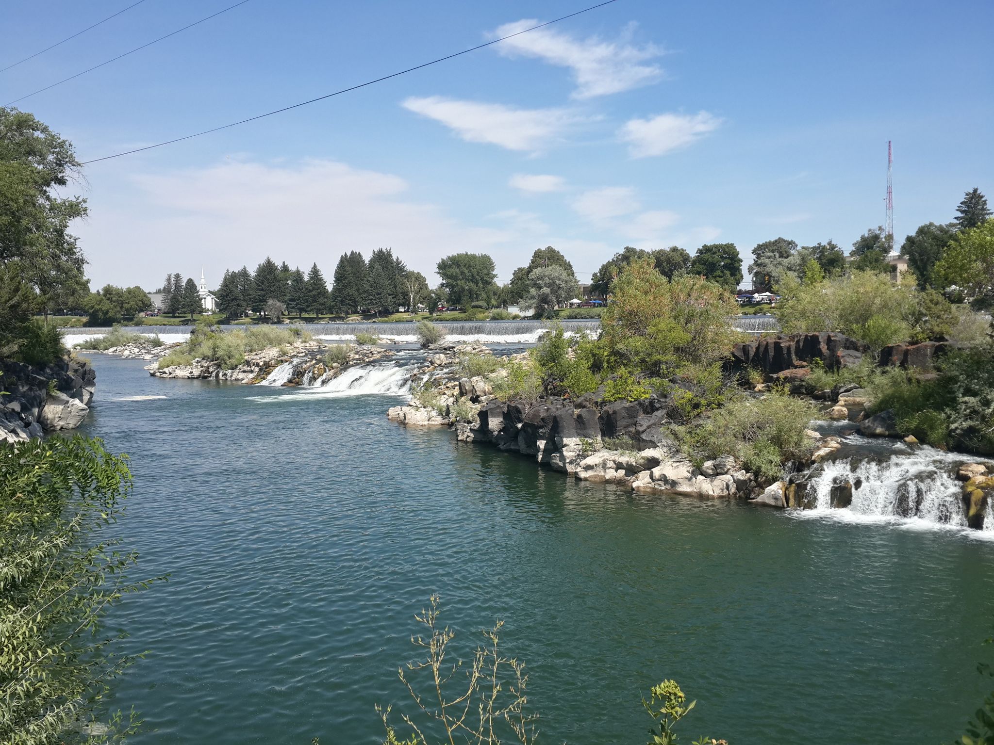 Wide river with small waterfalls, rocky islands, and green trees on a sunny day. - Idaho Falls Greenbelt loop