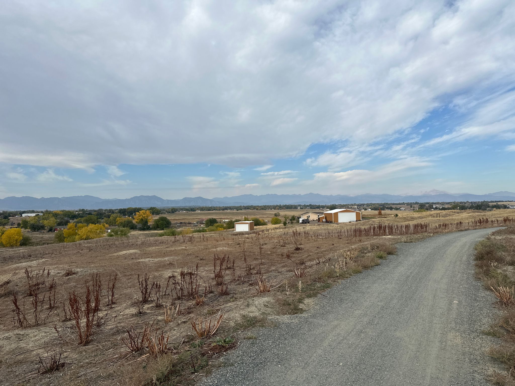 A gravel path winds through dry fields with distant buildings and mountains. - Sterns Lake – View of Longs Peak loop from Wetland Preserve