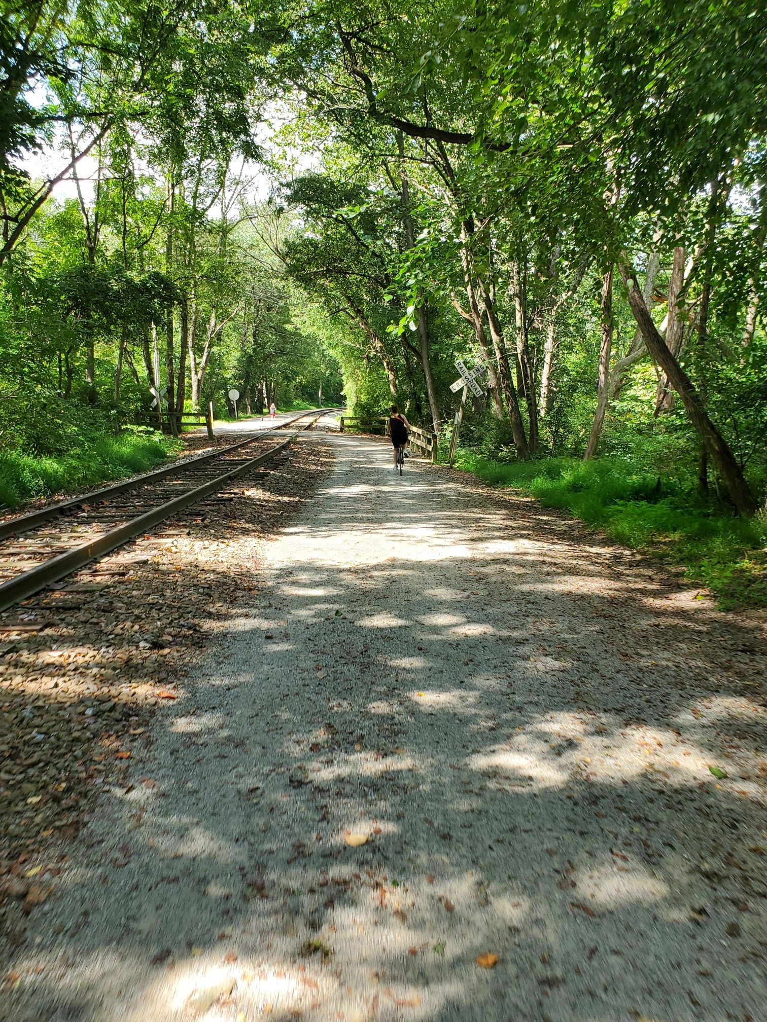 A person cycles on a sun-dappled gravel path beside railroad tracks. - York County Heritage Rail Trail
