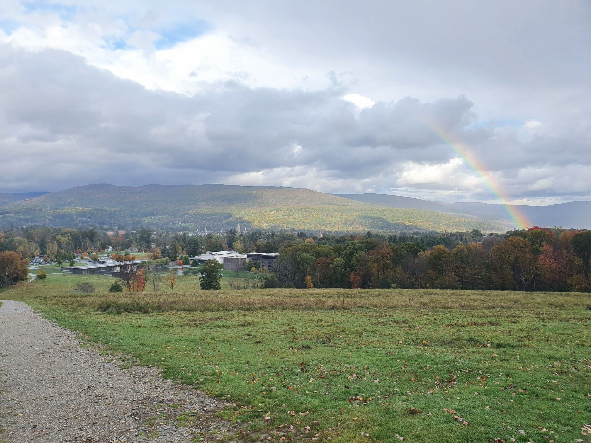 A rainbow arches over distant mountains and buildings on a cloudy day. - Fitch Memorial Lands – 1753 House loop from The Clark Art Institute