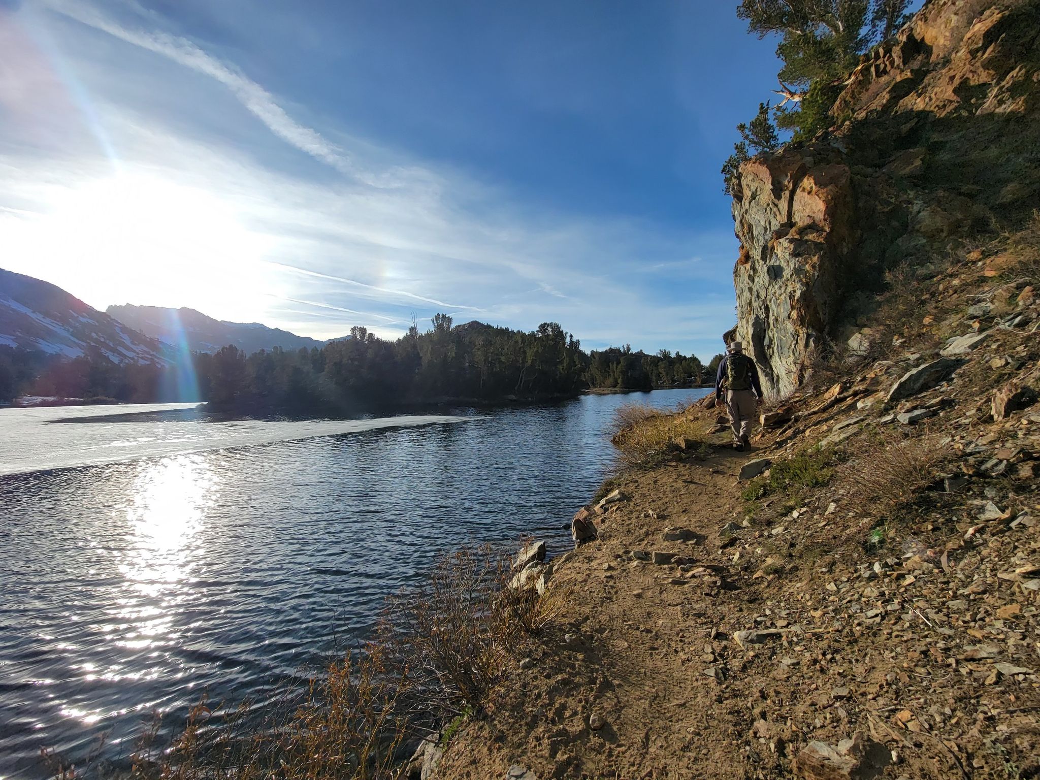Person walks along a dirt path beside a lake with sun glare. - Chocolate Lakes loop