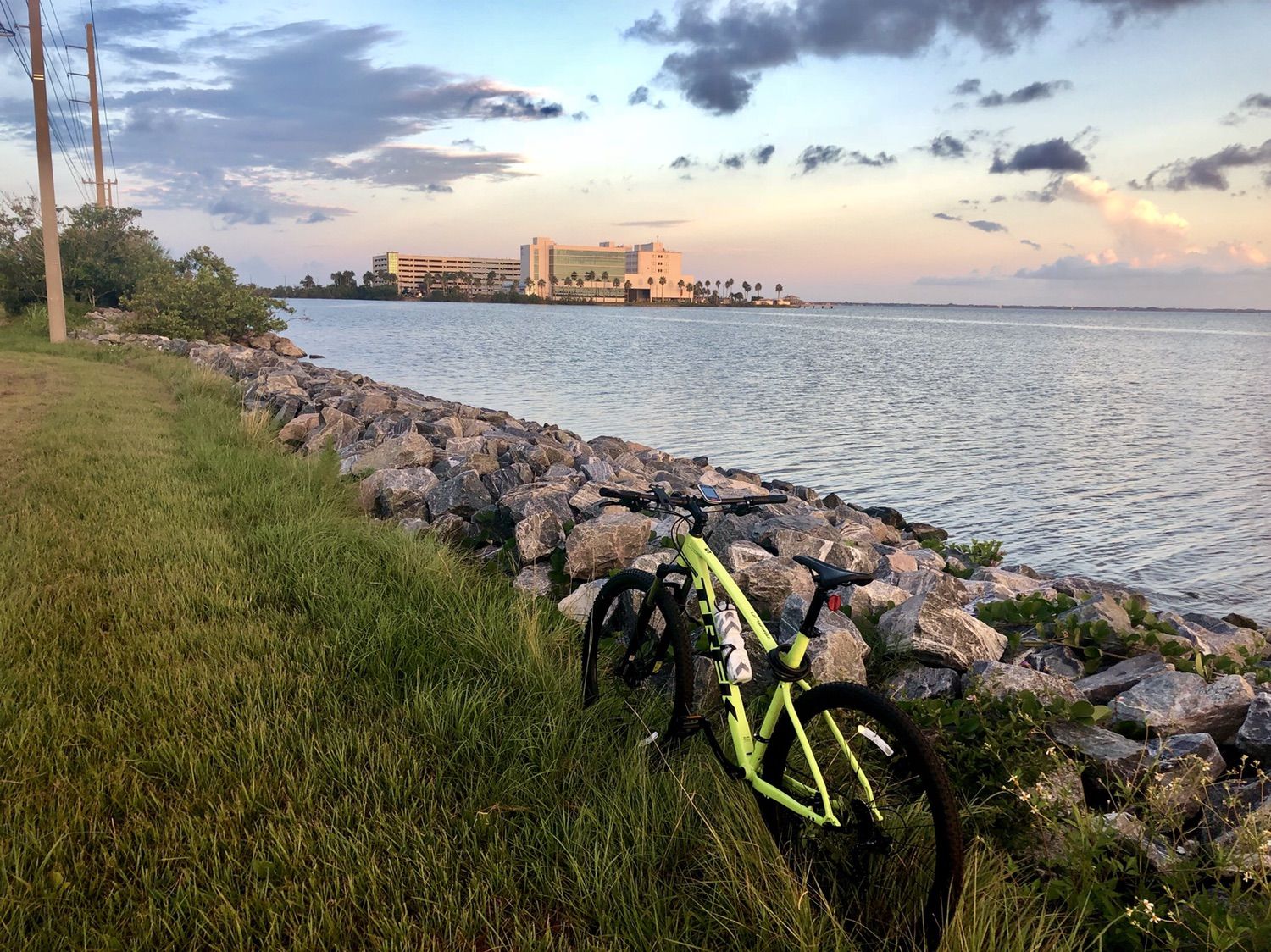Bright green bicycle rests by a rocky shore with calm water and distant buildings. - Merritt Island loop via Satellite Beach