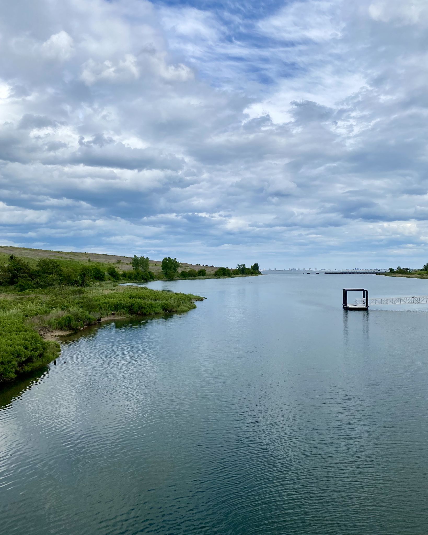 Calm water body with green banks and distant structures under cloudy sky. - Jamaica Bay Greenway Loop