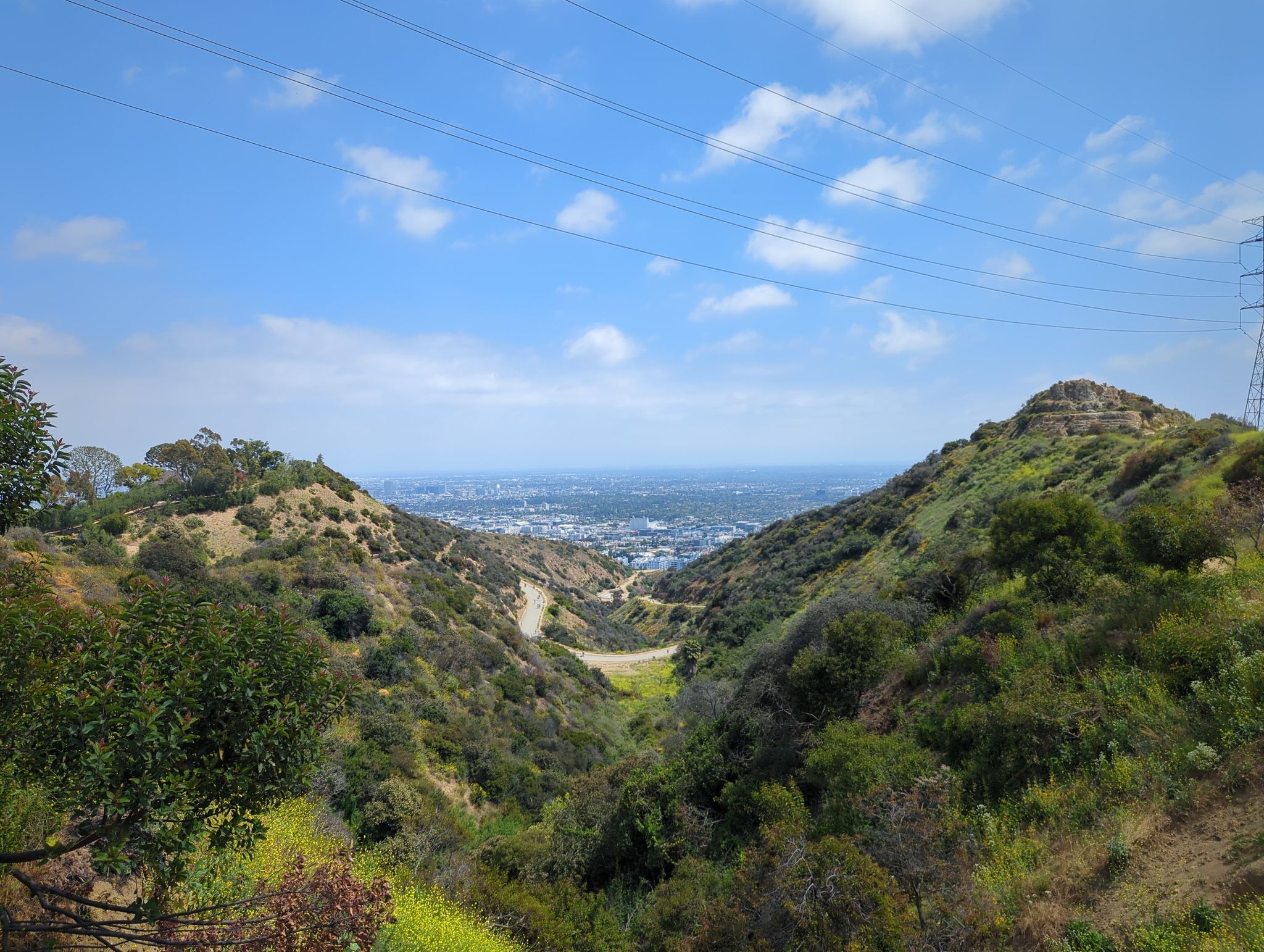 Green hills frame a distant city view under a bright blue sky with power lines. - Runyon Canyon Park loop from Hollywood Curson
