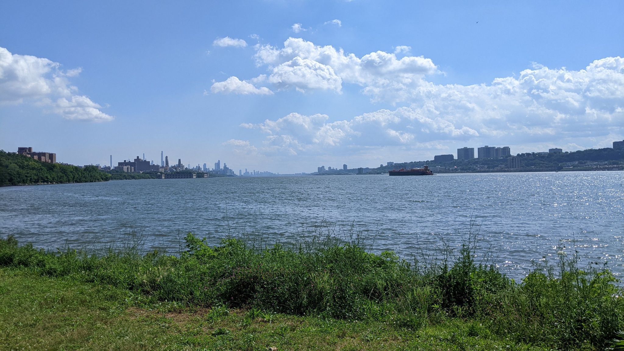 A wide river flows under a bright blue sky with distant cityscapes. - One World Trade Center – Hudson River Greenway loop from 215th Street