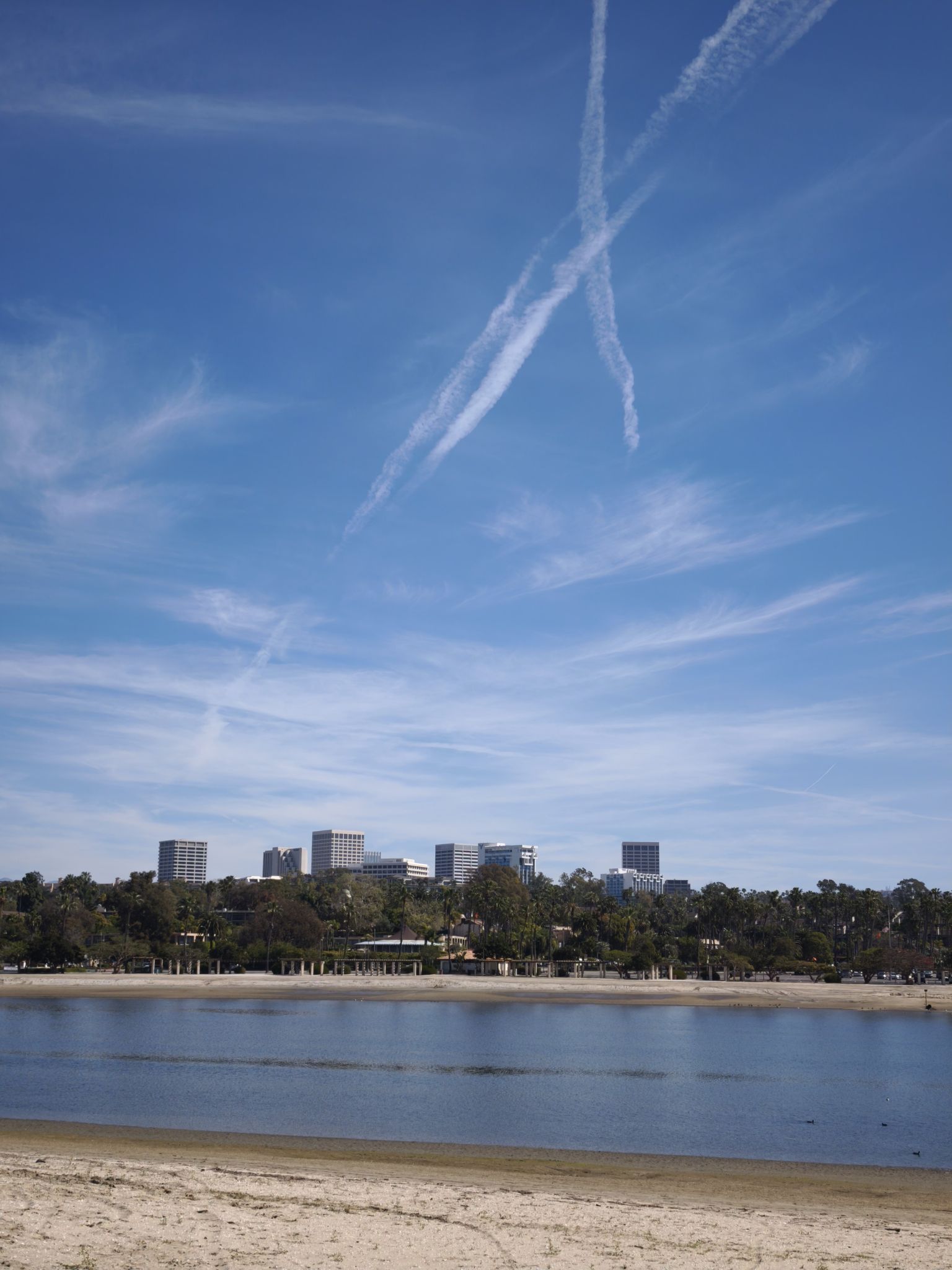 City skyline across water under a bright blue sky with contrails. - Newport Dunes Loop
