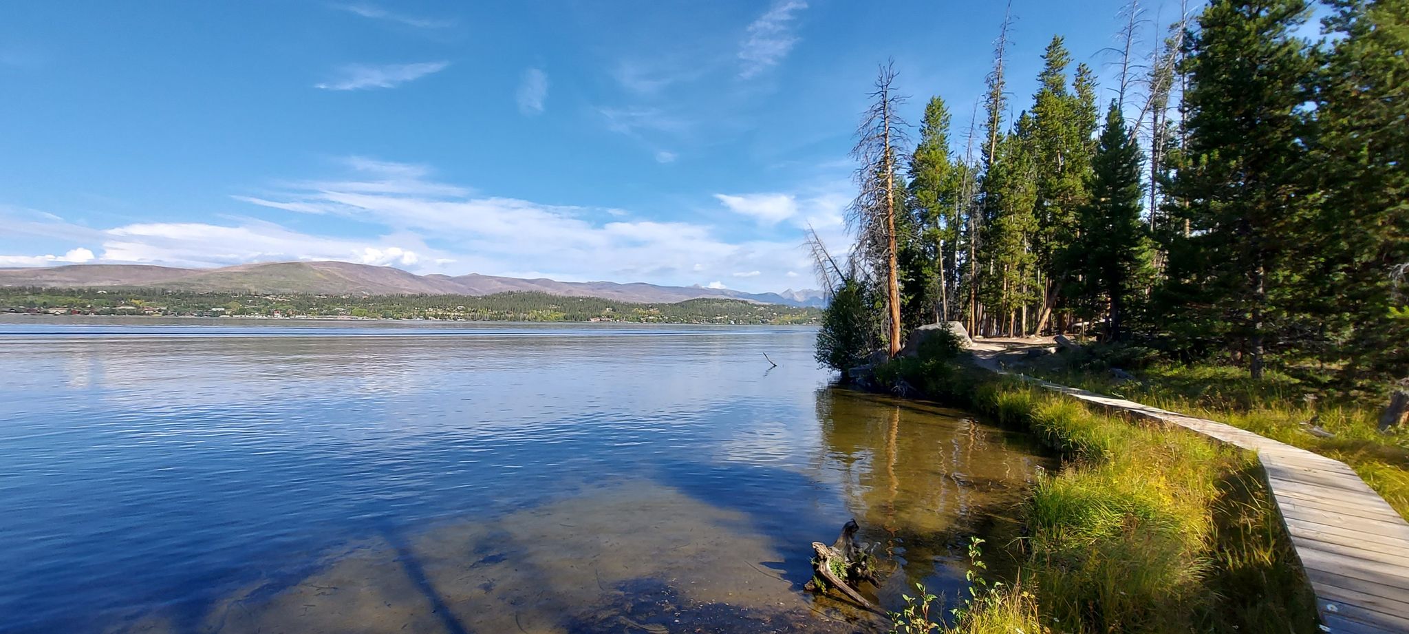 A wooden boardwalk follows the shore of a calm blue lake under a sunny sky. - East Shore Trail