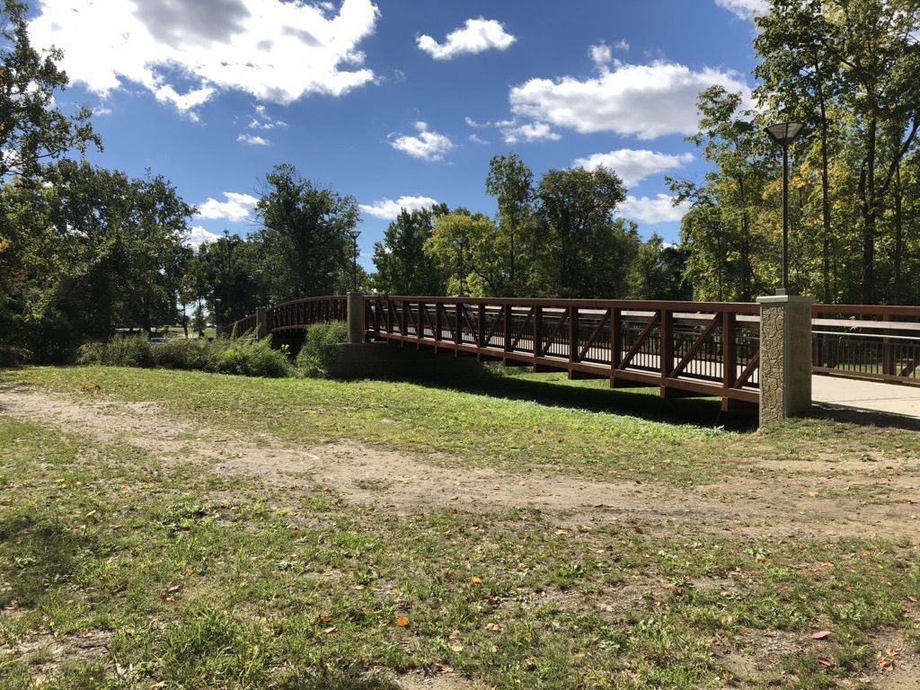 A long wooden pedestrian bridge spans a grassy area on a sunny day. - Clinton River Bridge – Clinton River Park loop from Adlai E. Stevenson High School