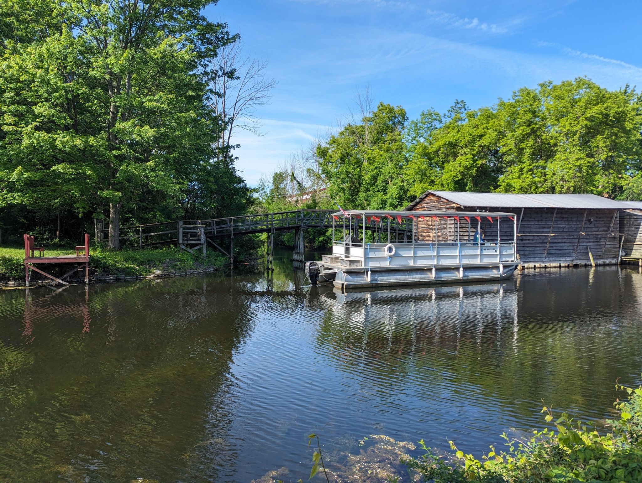 Pontoon boat docked by a wooden building and bridge over a calm waterway. - Nine Mile Creek Aqueduct – Erie Canal Park loop from Redman Cemetery