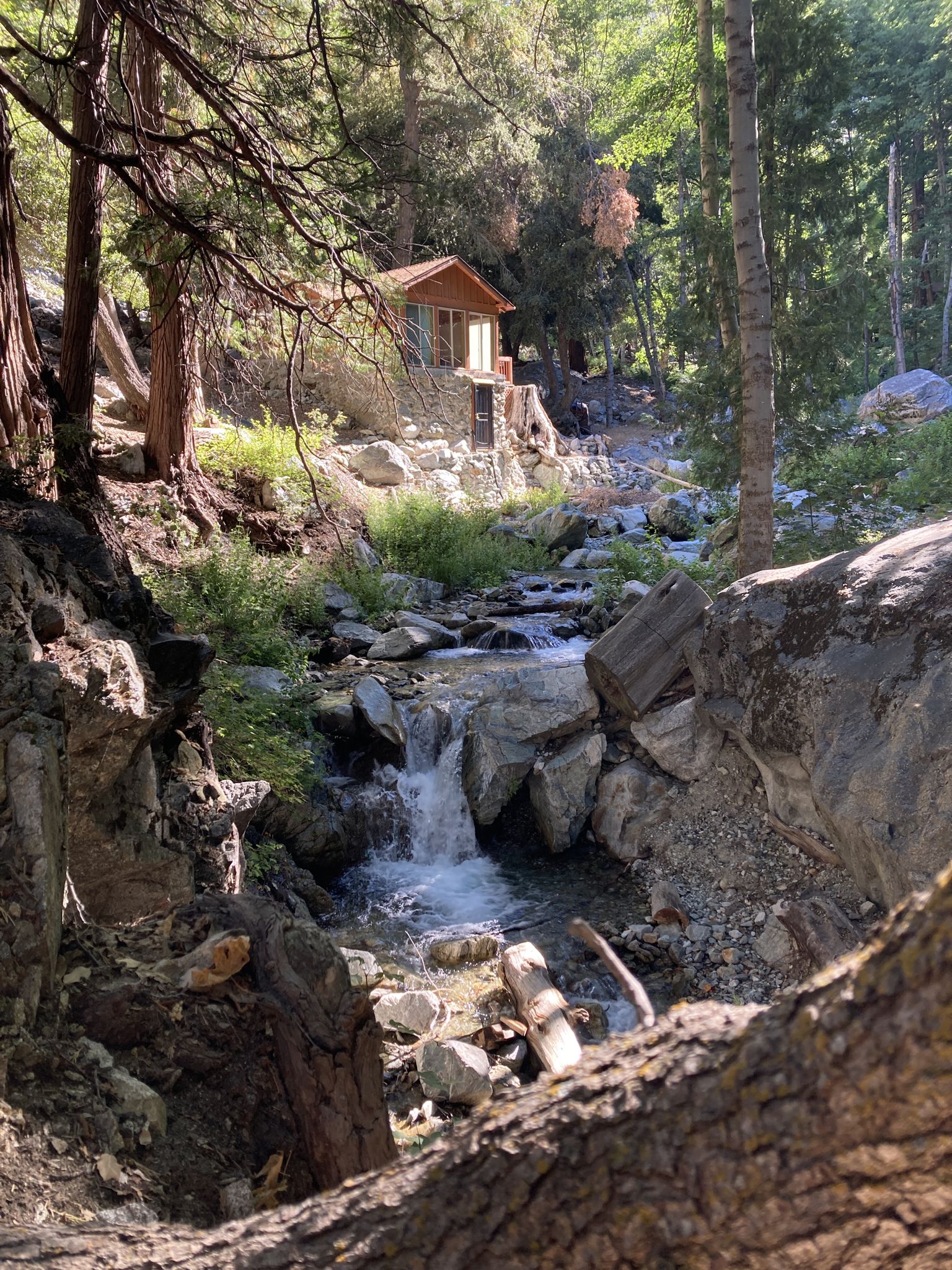 A small cabin overlooks a rocky stream with a waterfall in a sunny forest. - Icehouse Canyon Trail