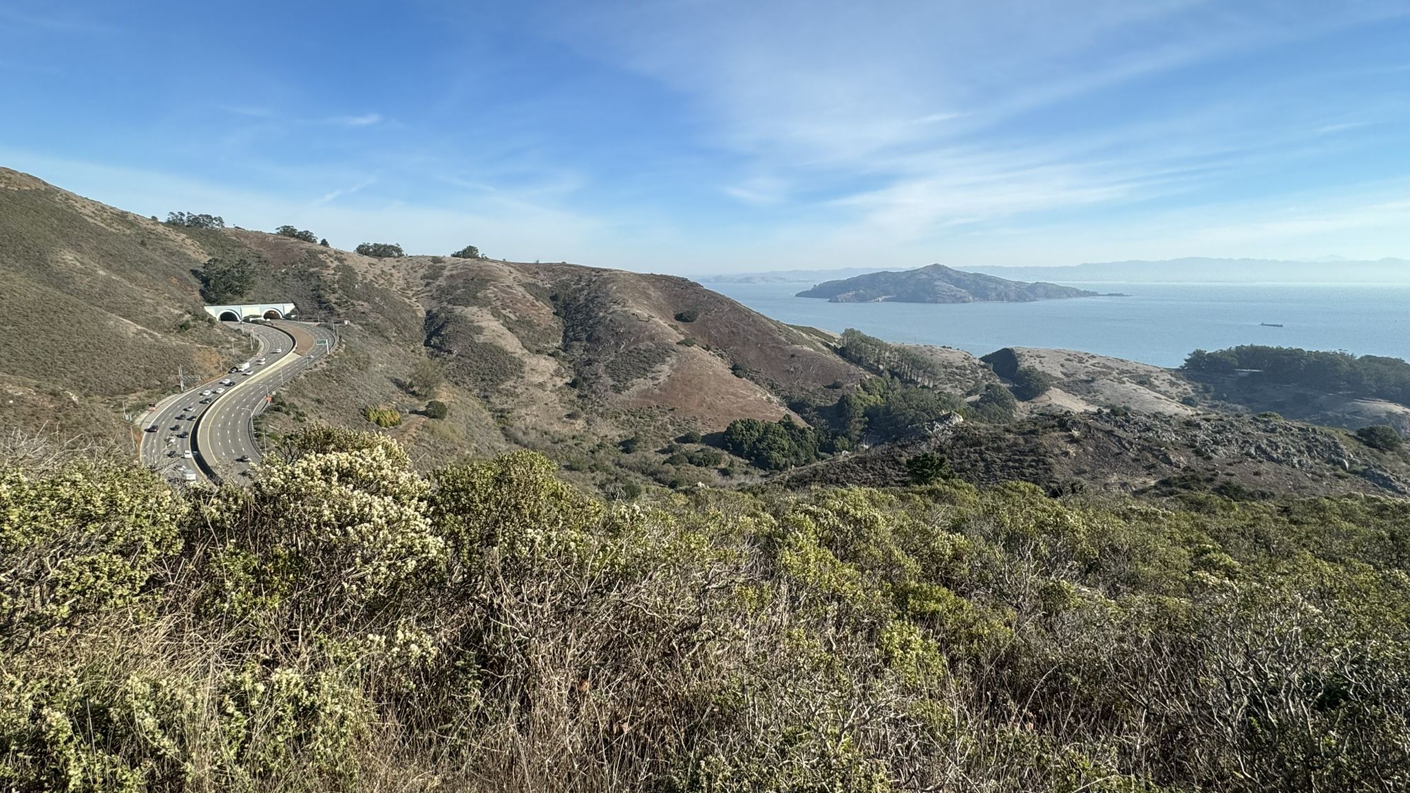 Sunny panoramic view of coastal hills, winding road, distant island, and ocean. - Slacker Hill and Hawk Hill via Coastal Trail