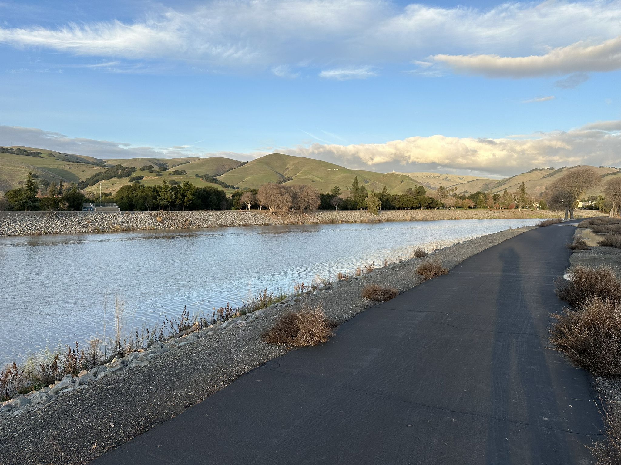 Paved path alongside calm water with rolling green hills under a partly cloudy sky. - Garin Regional Park loop