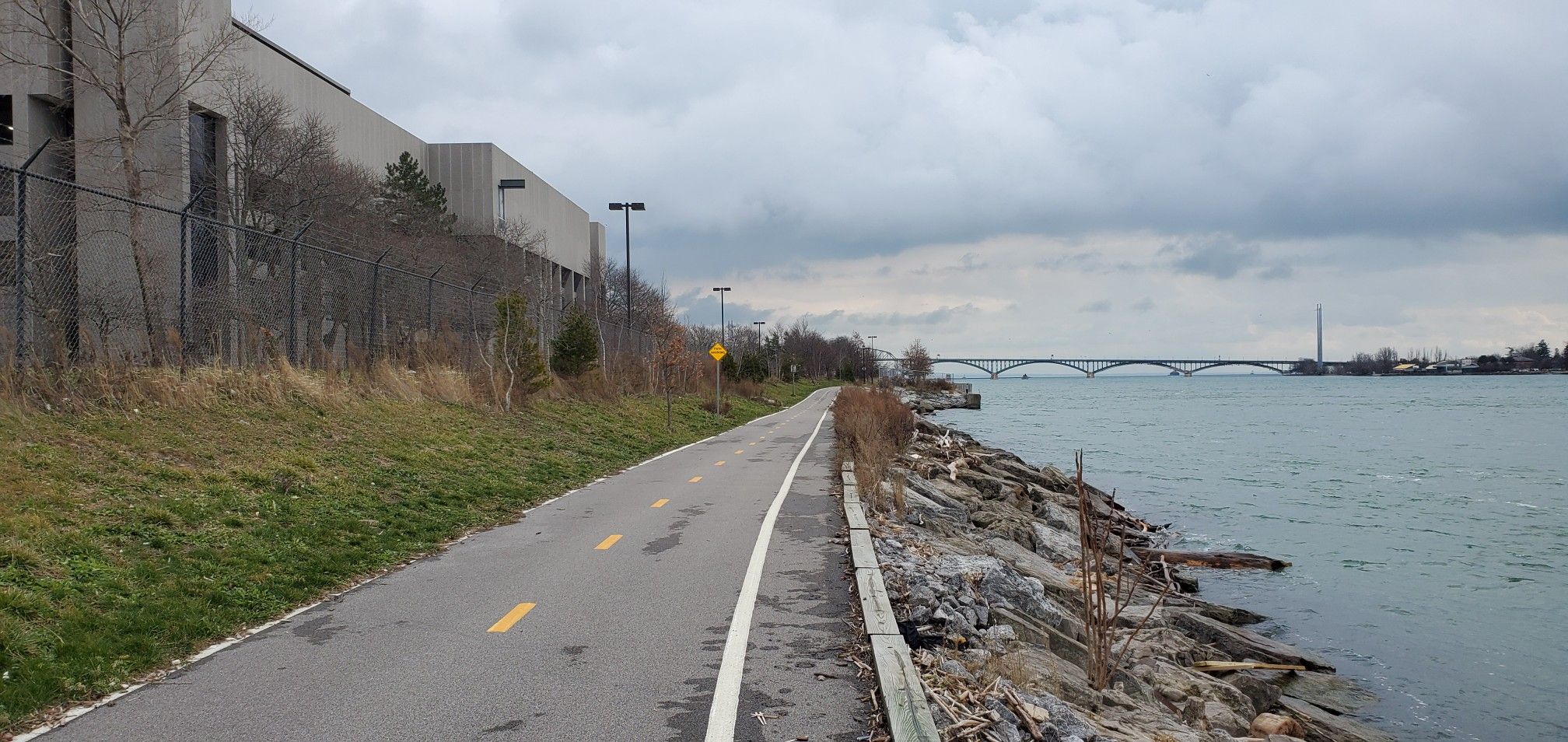Paved path beside a rocky shoreline and river, with a bridge in the distance. - Tonawanda to Buffalo via Shoreline Trail Riverwalk
