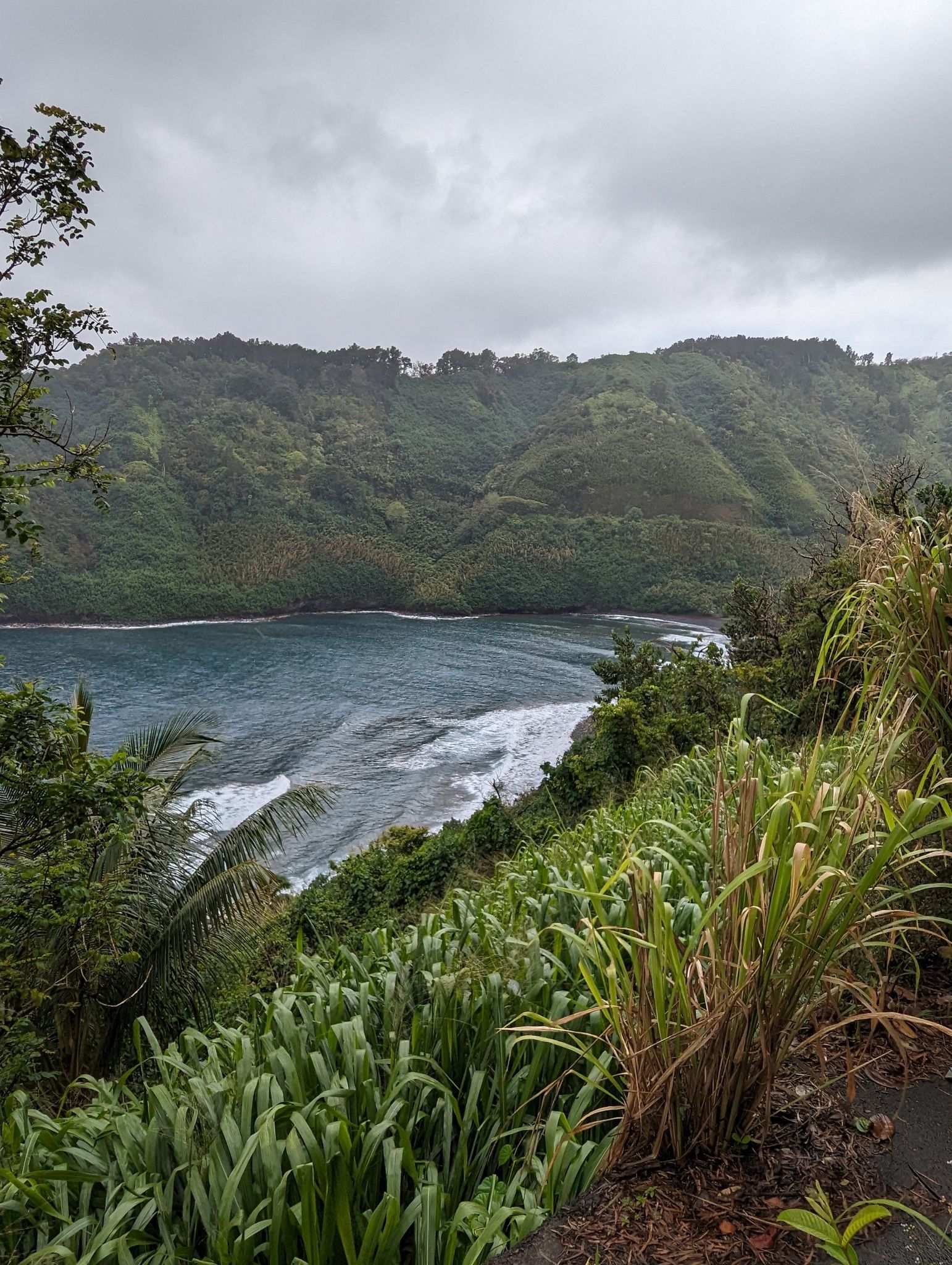 Overcast view of a lush green bay with crashing waves and steep hills. - Hāna Highway – Rodeo General Store loop from Maui