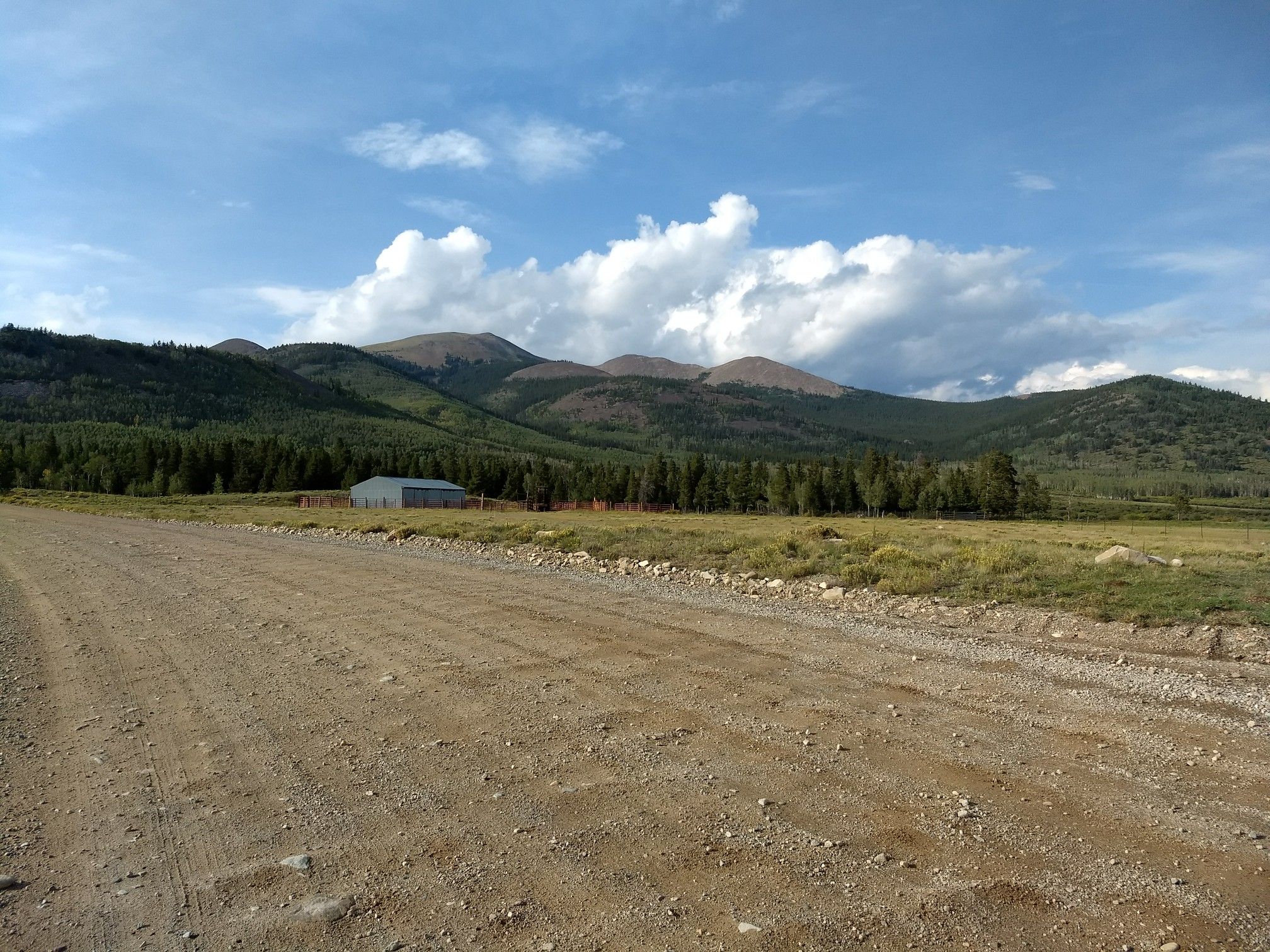 Dirt road leads to a building, forest, and mountains under a cloudy sky. - Boreas Pass