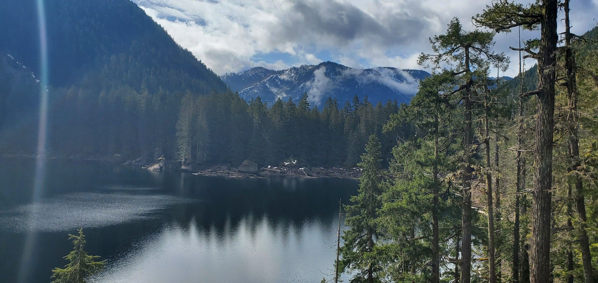 Sunlit lake, dense forest, and distant snow-capped mountains under a cloudy sky. - Upper Lena Lakes