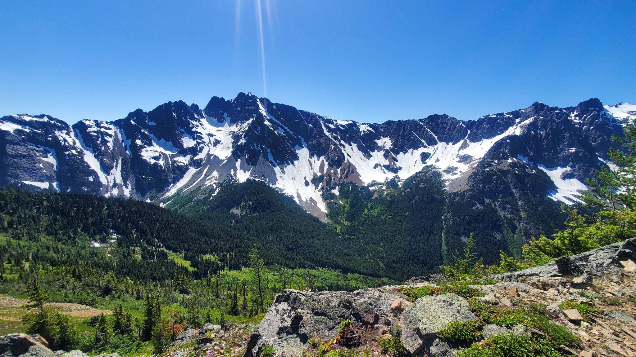 Sunny panoramic view of snow-capped mountains, green forests, and rocky foreground. - Grasshopper Pass via the PCT
