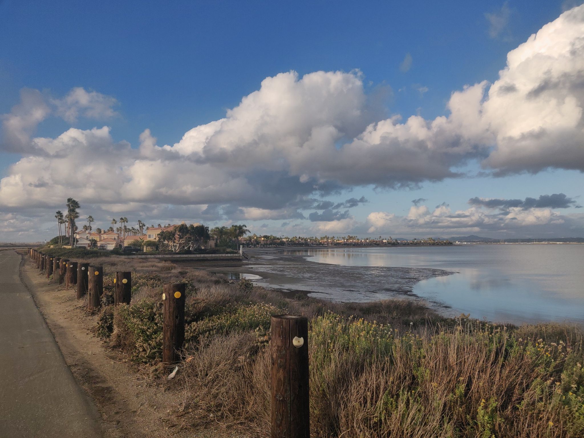 Dirt path with wooden posts beside calm water, distant buildings, cloudy sky. - Coronado Bayshore Bikeway – View of San Diego Bay loop from San Diego