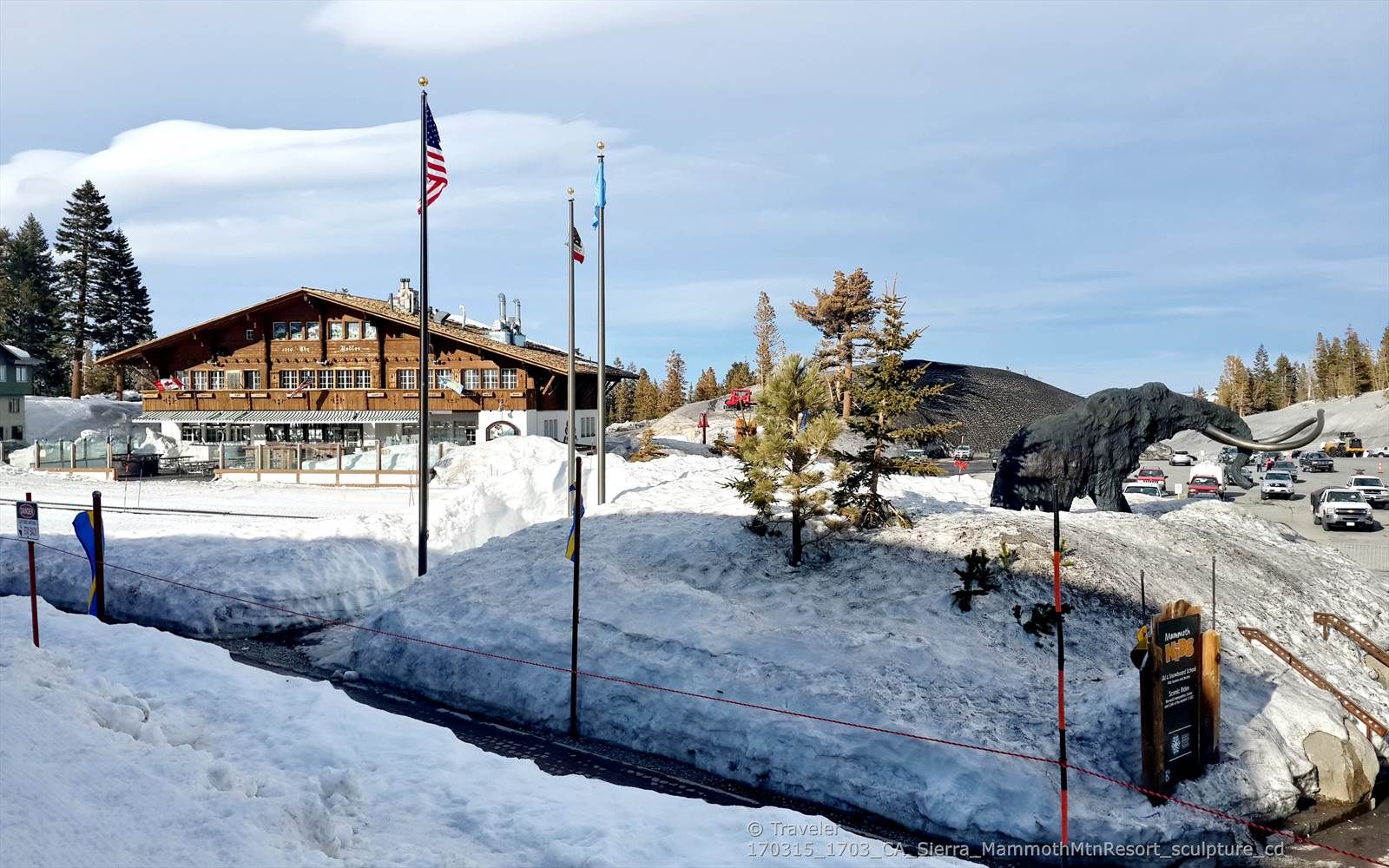 Snowy scene with a large building, flags, and a dark mammoth sculpture. - Village Plaza – Mammoth Mountain Resort loop from Eagle Lodge