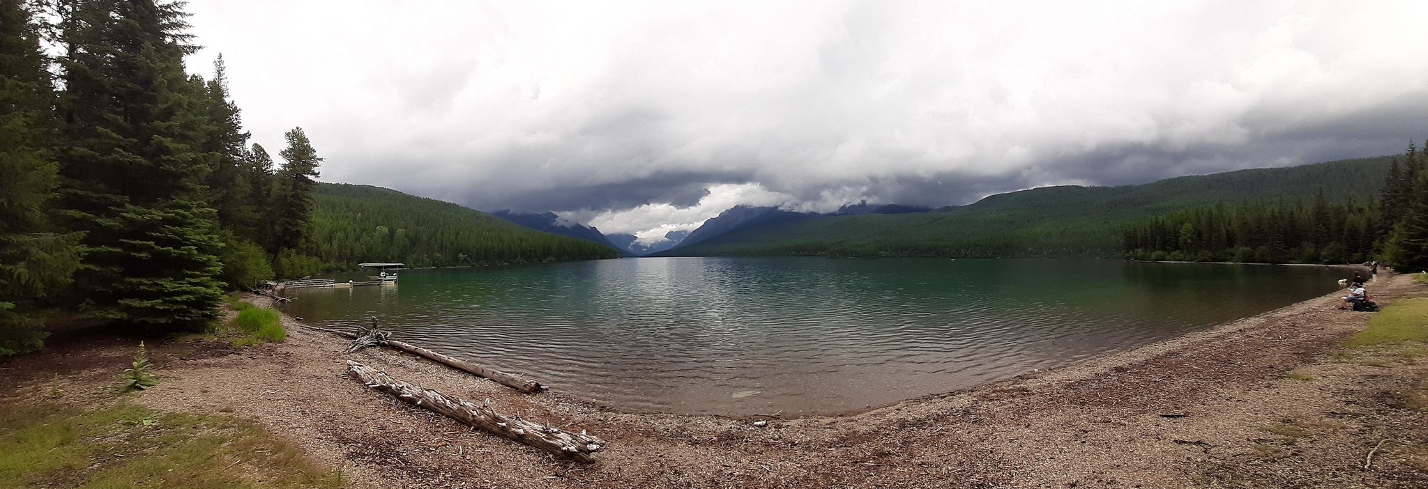 Panoramic view of a lake, forested shores, distant mountains, and two people relaxing. - Bowman Lake Trail