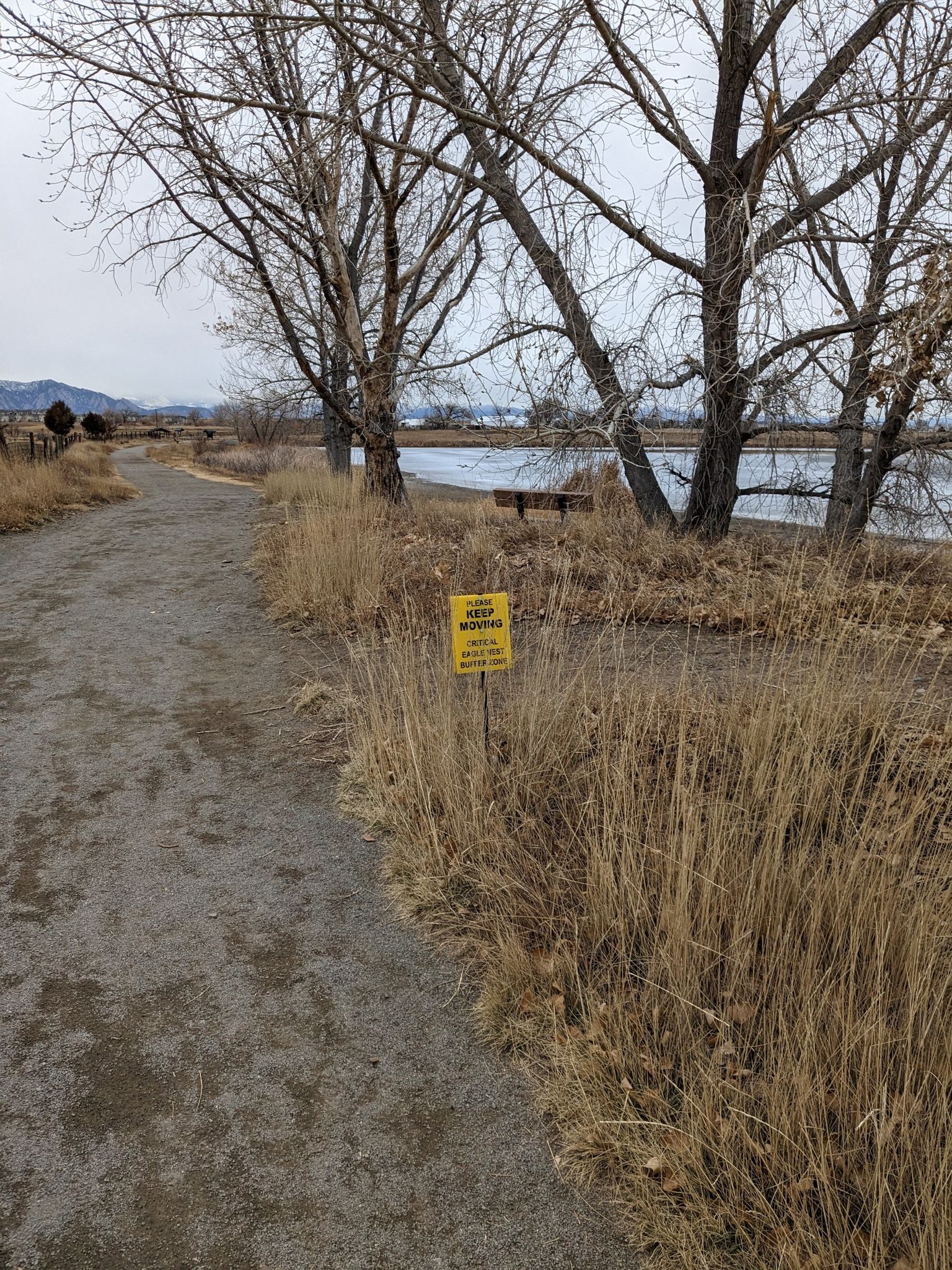 A dirt path next to dry grass, a yellow sign, and a lake. - Rock Creek Trail