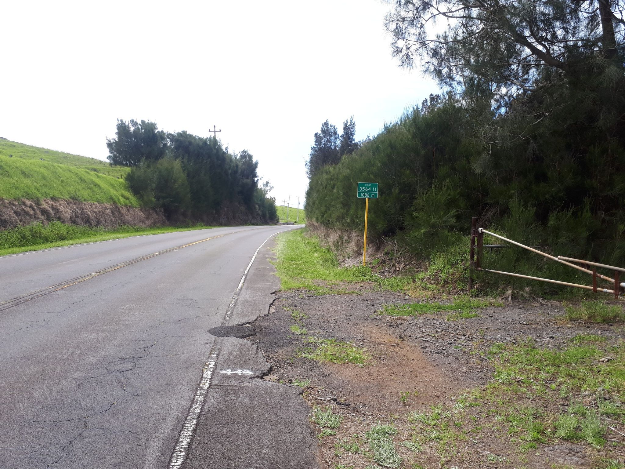 Paved road curves right next to a green sign and metal gate. - Kohala Mountain Road to Kahua Ranch