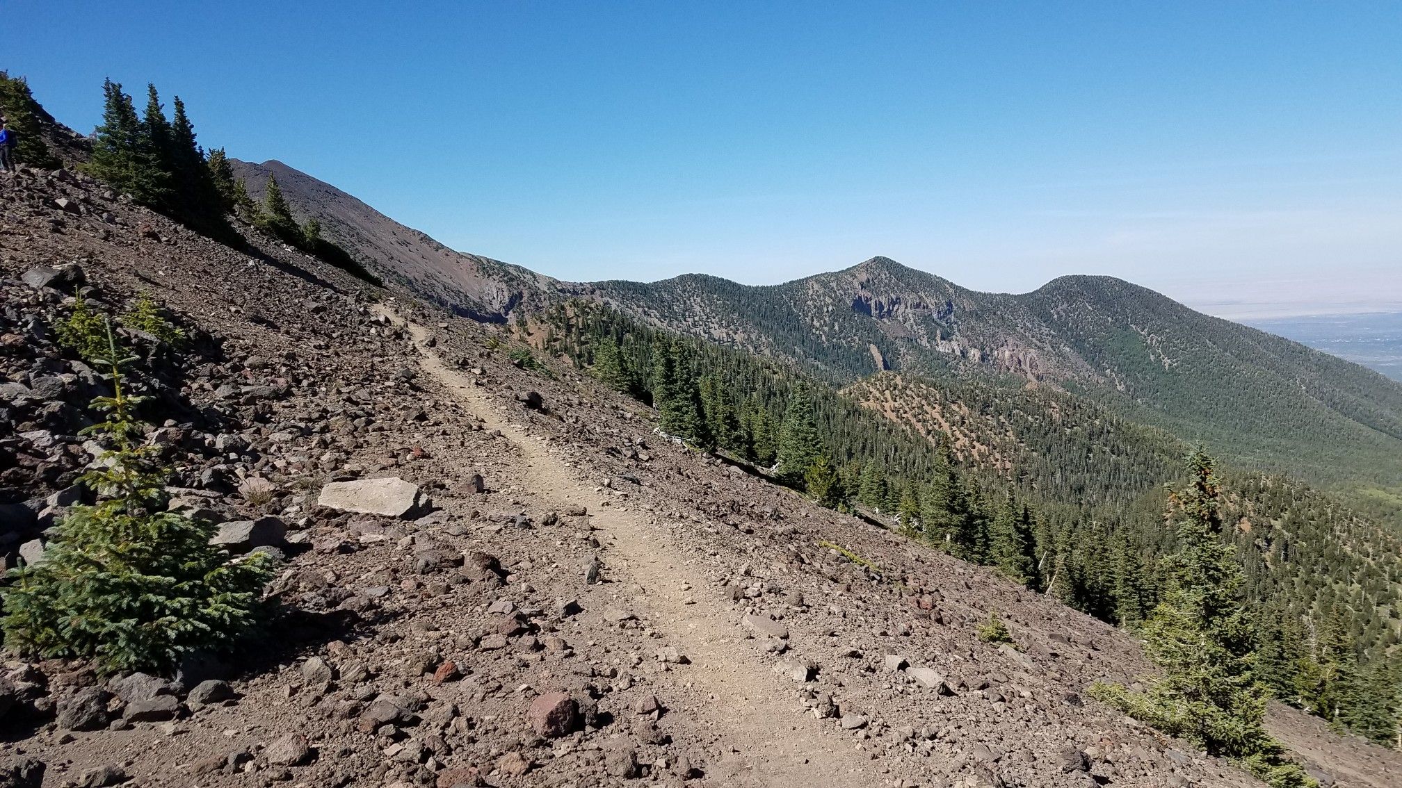 Narrow dirt trail traversing a steep, rocky mountain slope with distant forests. - Weatherford Trail