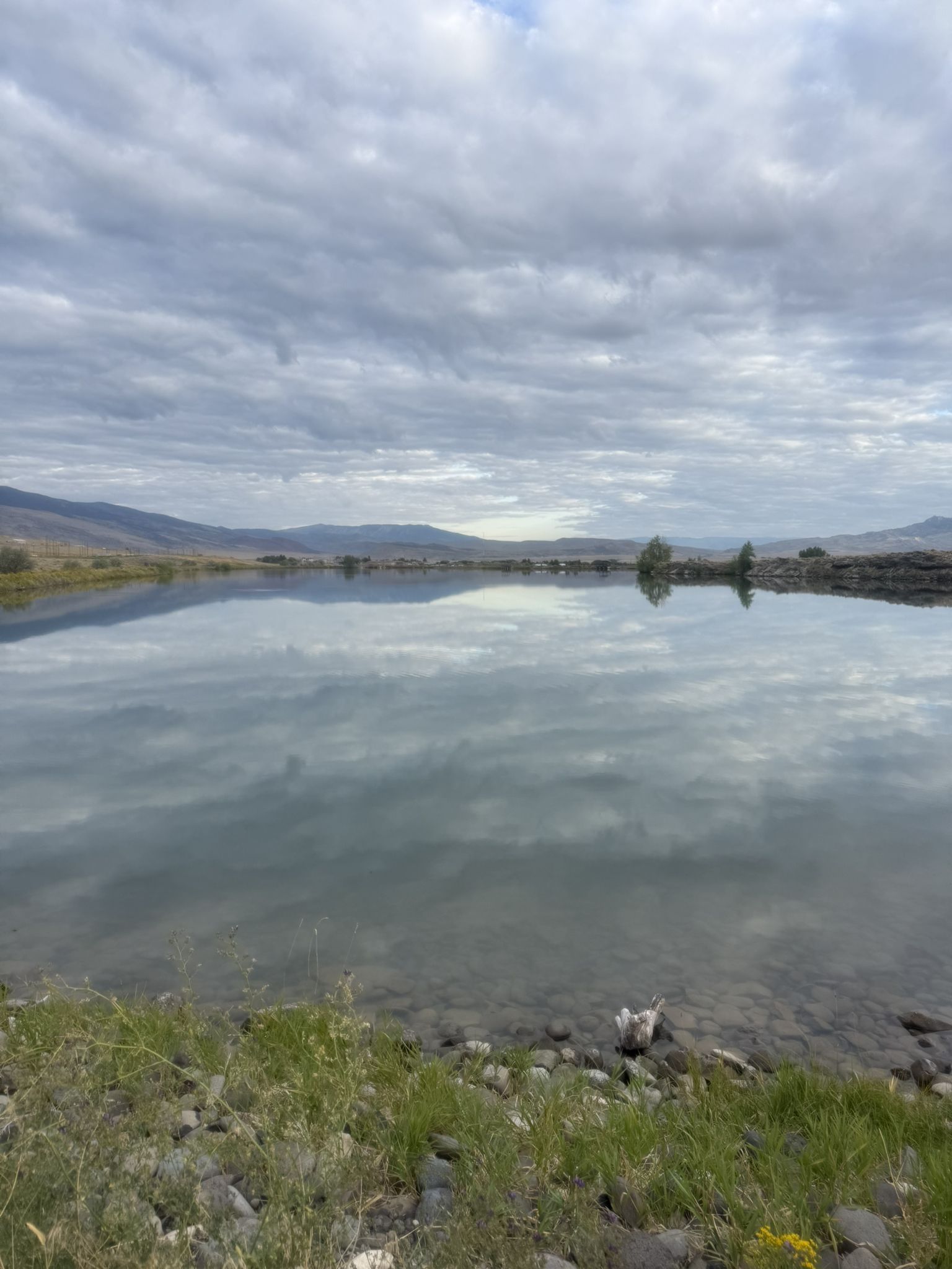 Calm lake reflecting cloudy sky, with distant mountains and rocky shore. - New Cody Reservoir loop from Cody
