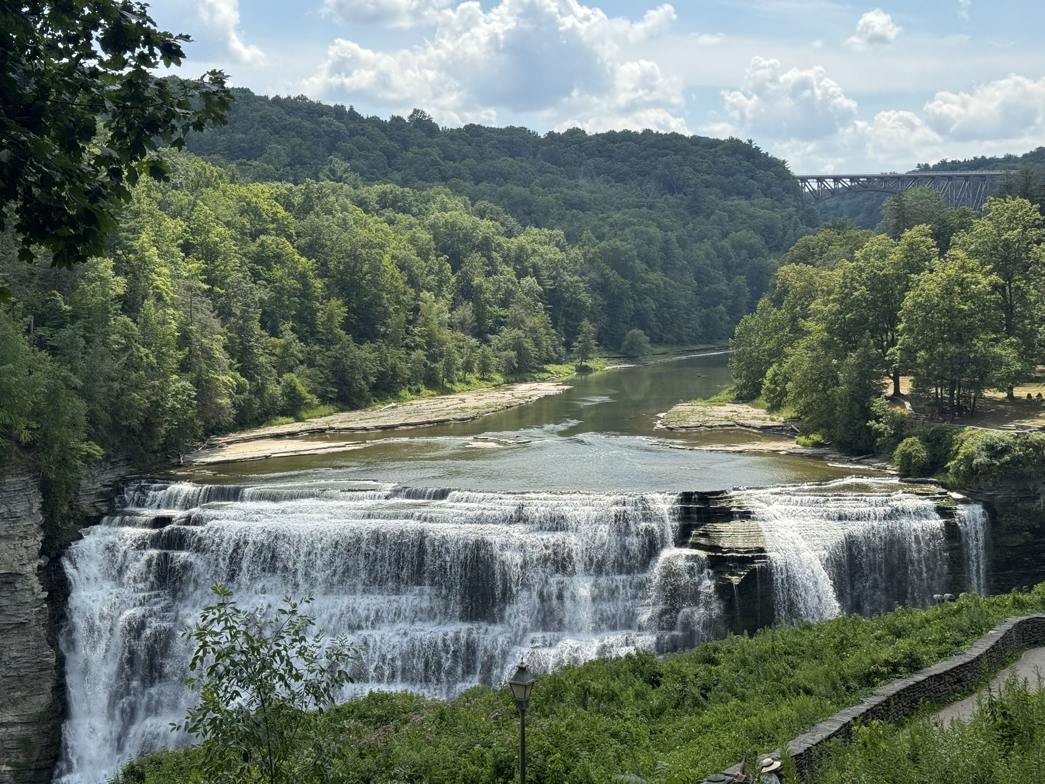 Wide waterfall flowing into a river, surrounded by green trees and a distant bridge. - Tea Table loop from Chipmunk Trail (#19A)