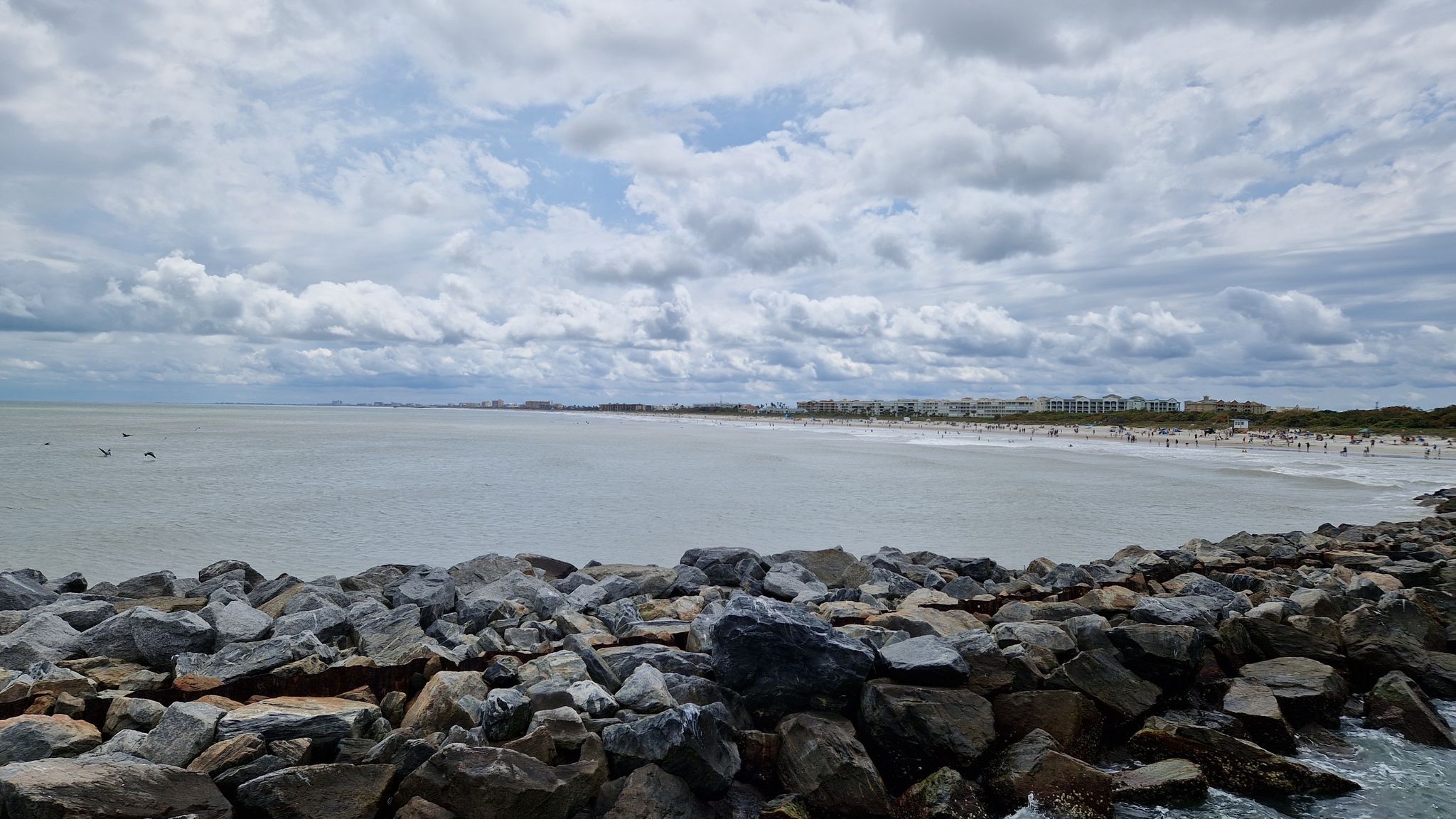 Wide view of a cloudy beach with ocean, rocks, and distant people. - Hiking loop from Cape Canaveral