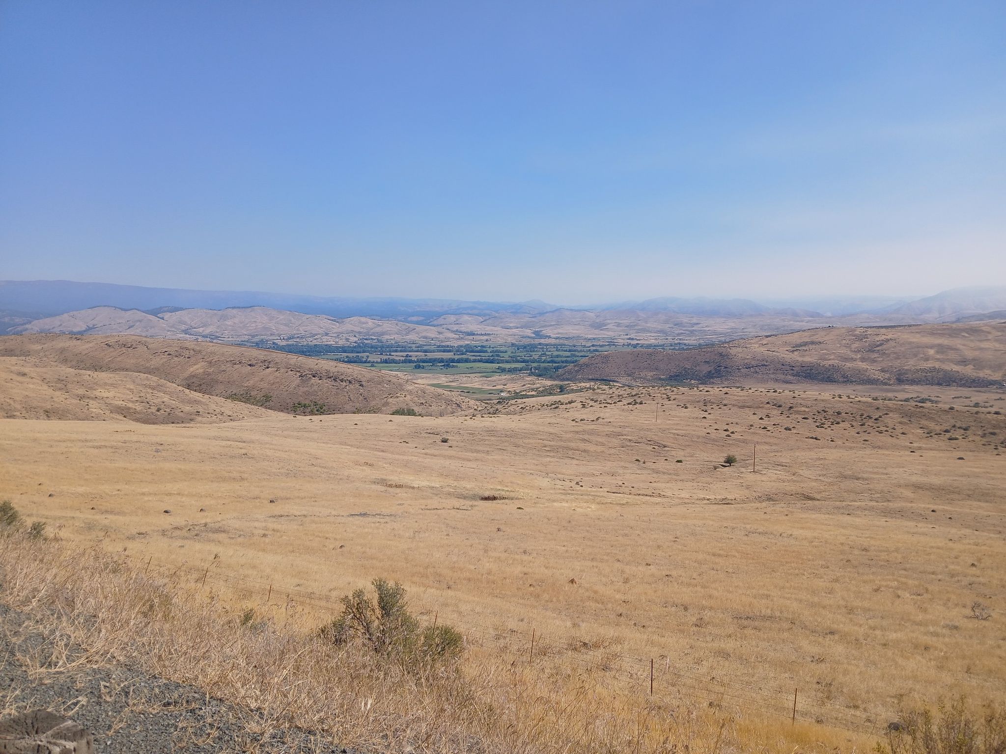 Golden, dry hills overlooking a green valley under a clear, hazy sky. - Halfway from Baker City via Hells Canyon Scenic Byway