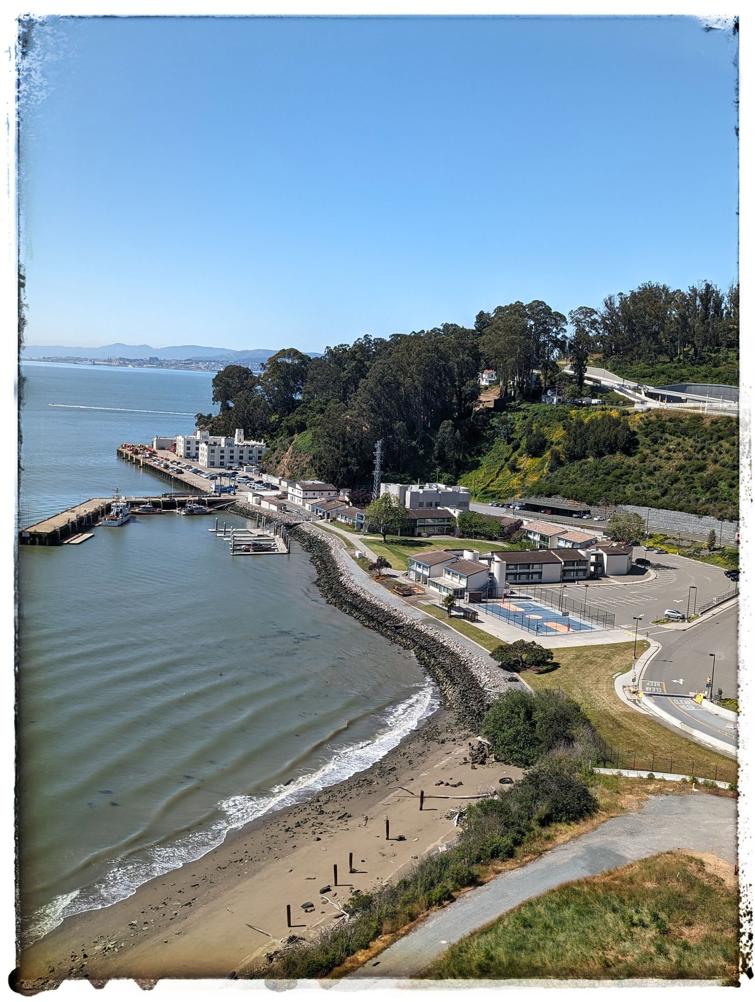 Aerial view of a bay with a beach, buildings, and a road winding up a hill. Sunny. - Bridge Yard – Emeryville Bayfront Bridge loop from Emeryville