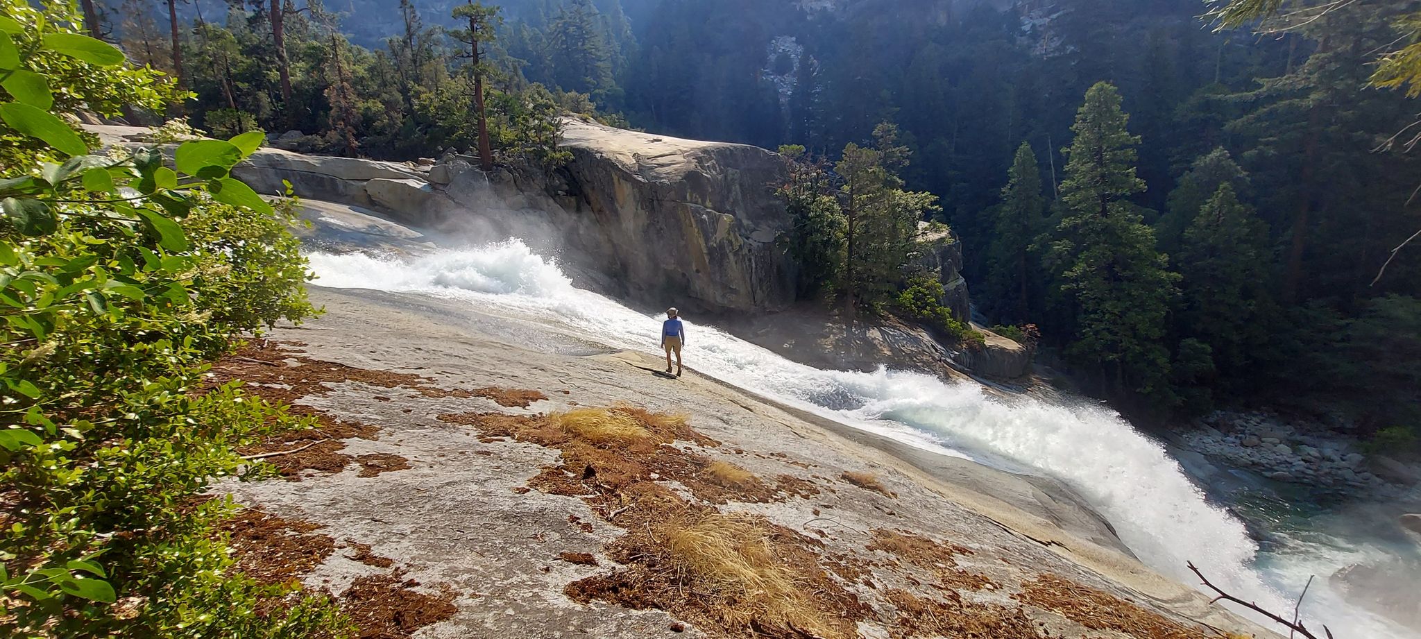Person standing on a large rock slab next to a powerful waterfall, sunny. - Mist Falls