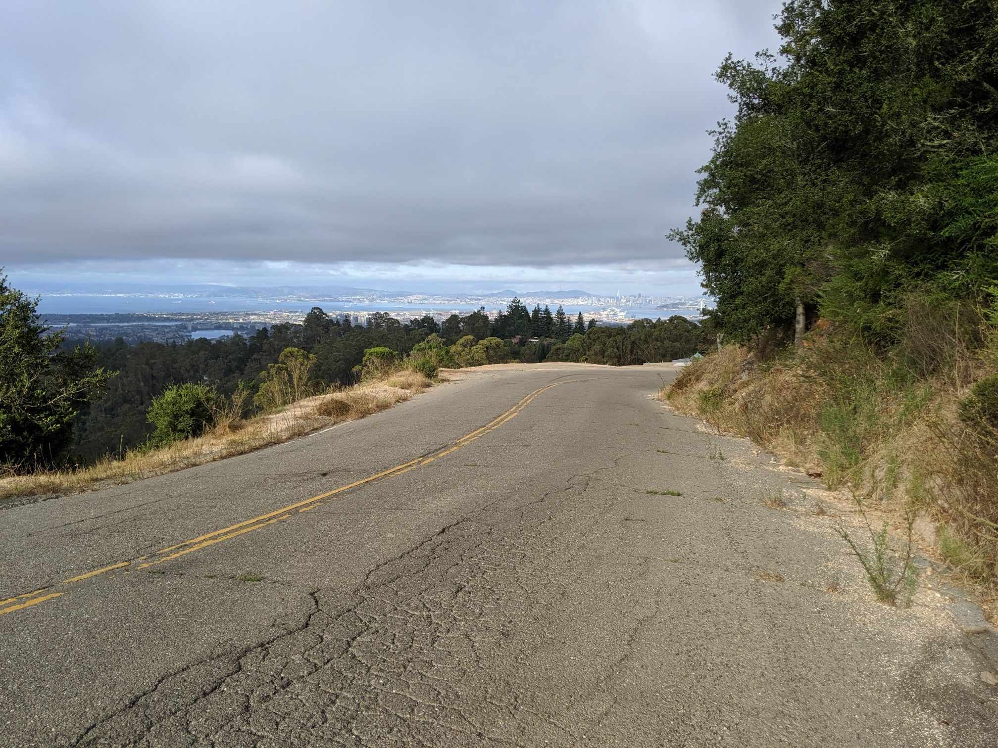 Empty cracked asphalt road winding through green hills under a cloudy sky. - Skyline Boulevard – Joaquin Miller Park loop from Ashby