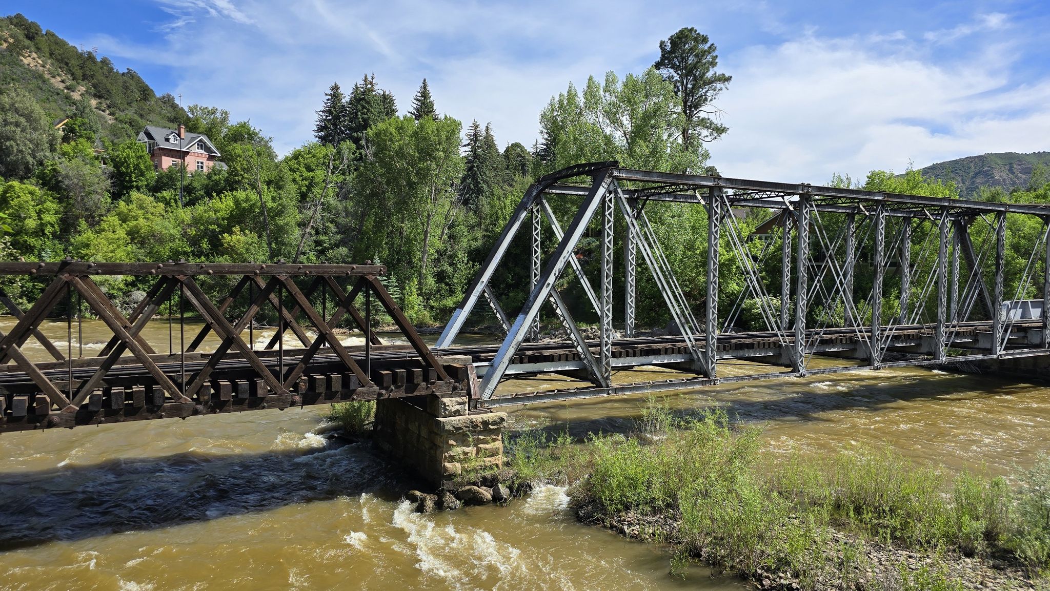 Two metal truss bridges span a muddy river under a bright sky. - Animas River Trail