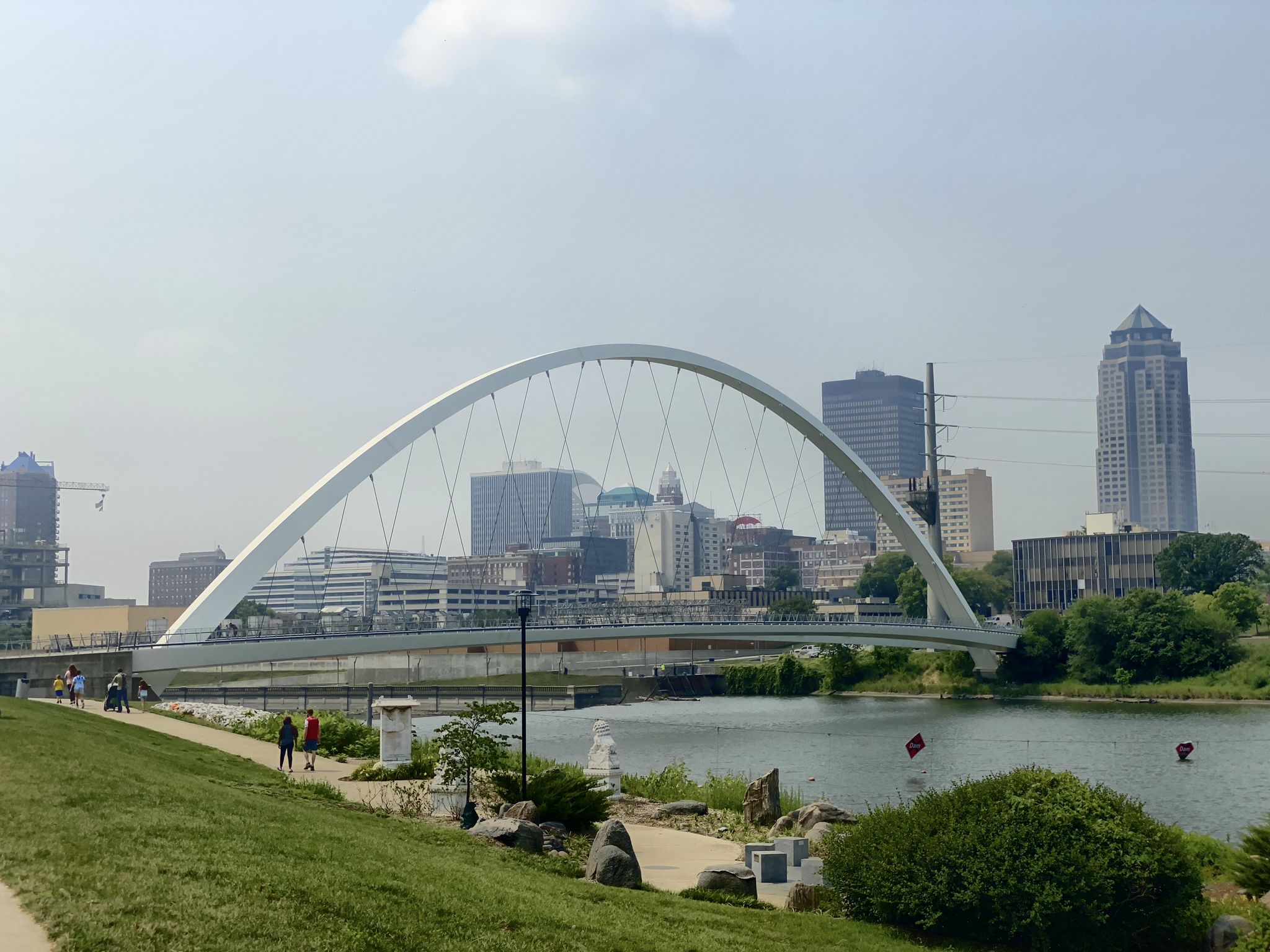 People walk near a modern arched bridge and city skyline over a river. - Gray's Lake beach – Principal Riverwalk loop from Terrace Hill