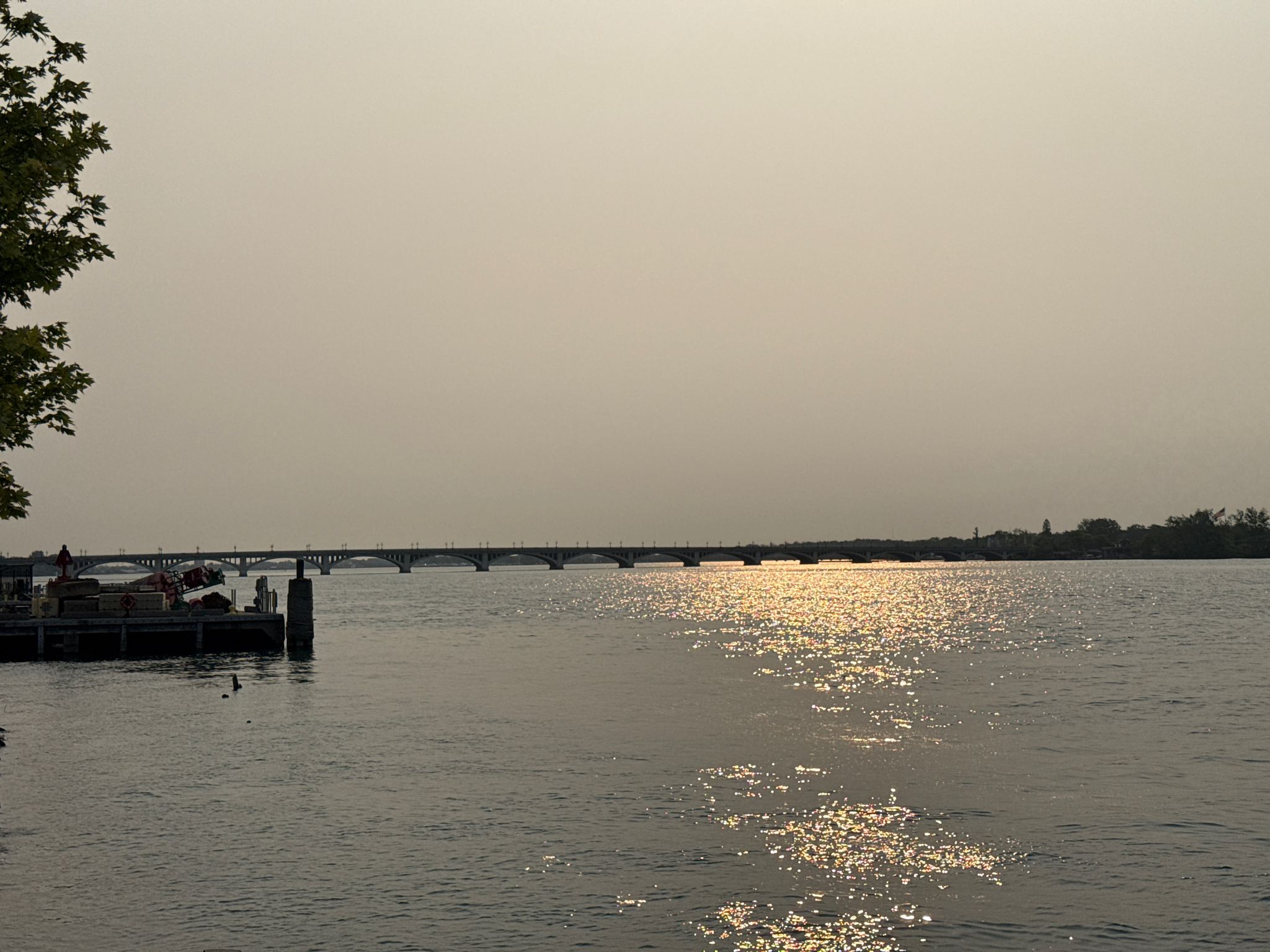 Hazy sunset over water with a distant bridge and figures on a dock. - Detroit Riverwalk and Dequindre Cut Loop