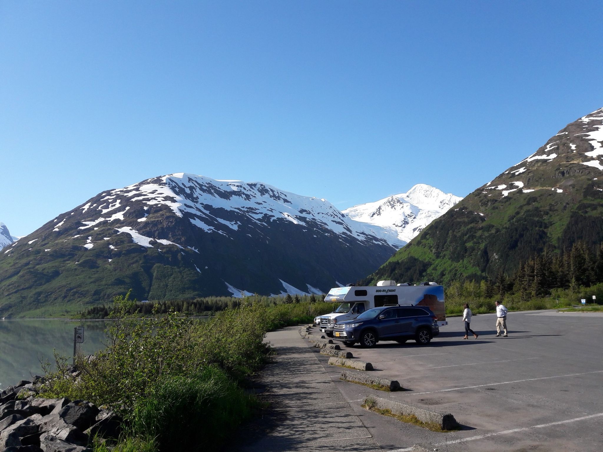 An RV and car parked by a lake, with snow-capped mountains and people. - Trail of Blue Ice