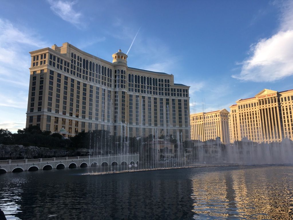 Grand building with fountains spraying water in a lake under a blue sky. - Excalibur Hotel and Casino – Fountains of Bellagio loop from MGM Grand Garden Arena