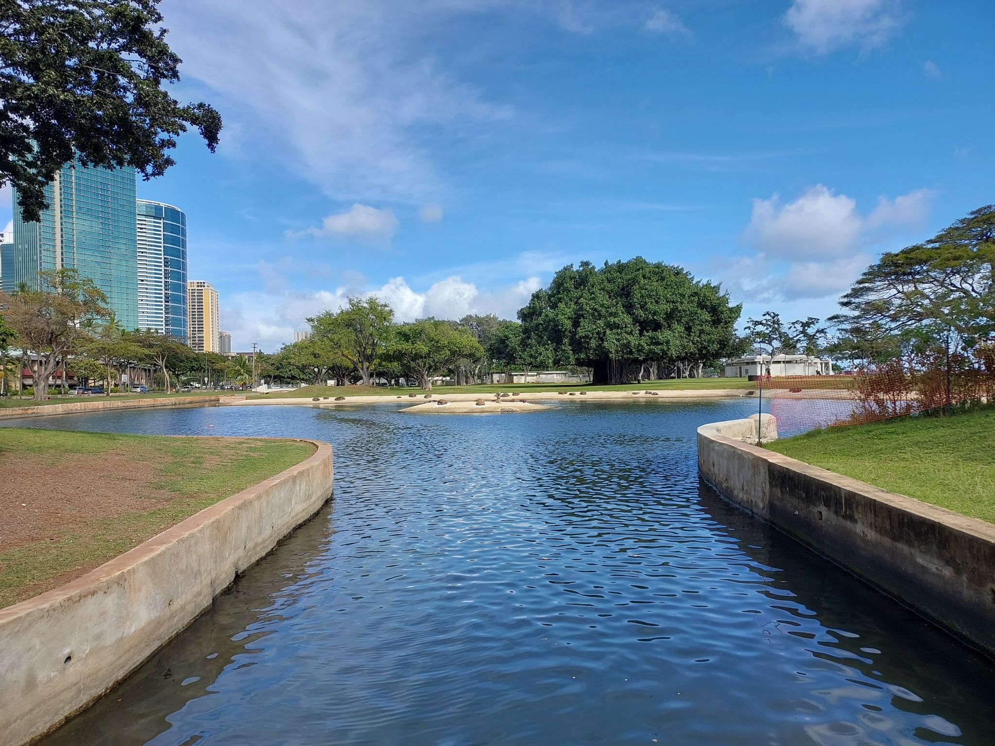 Calm canal water with green banks, large trees, and city buildings. - Lei of Parks Trail