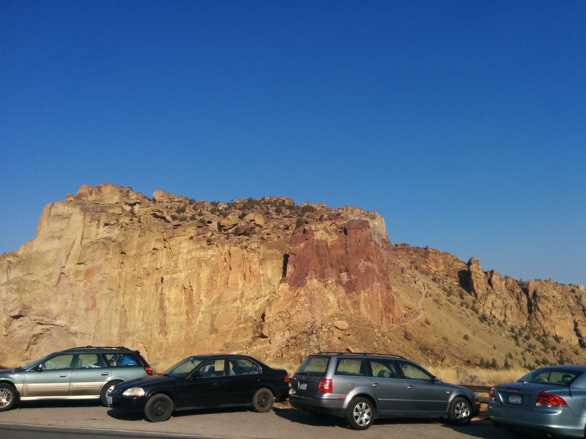 Several cars parked along a road with a massive rocky cliff in background. - Sisters to Smith Rock and Twin Bridges Loop