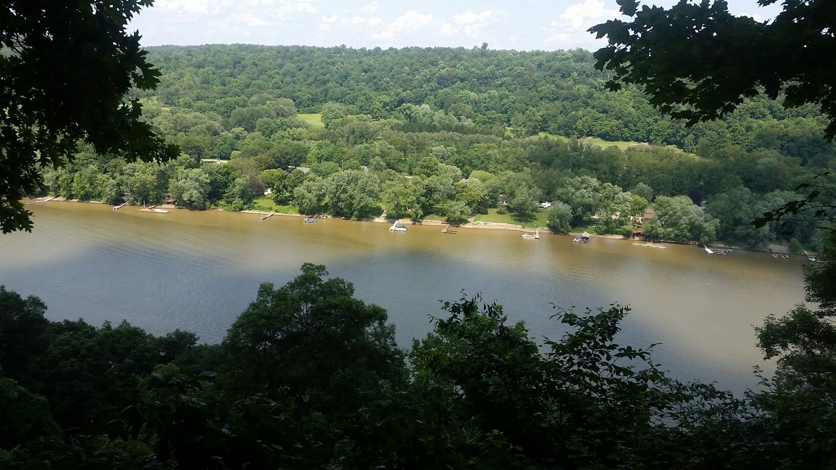 Wide river flowing through a dense green forest under a partly cloudy sky. - Woodlands, Wetlands, and Scouts Loop